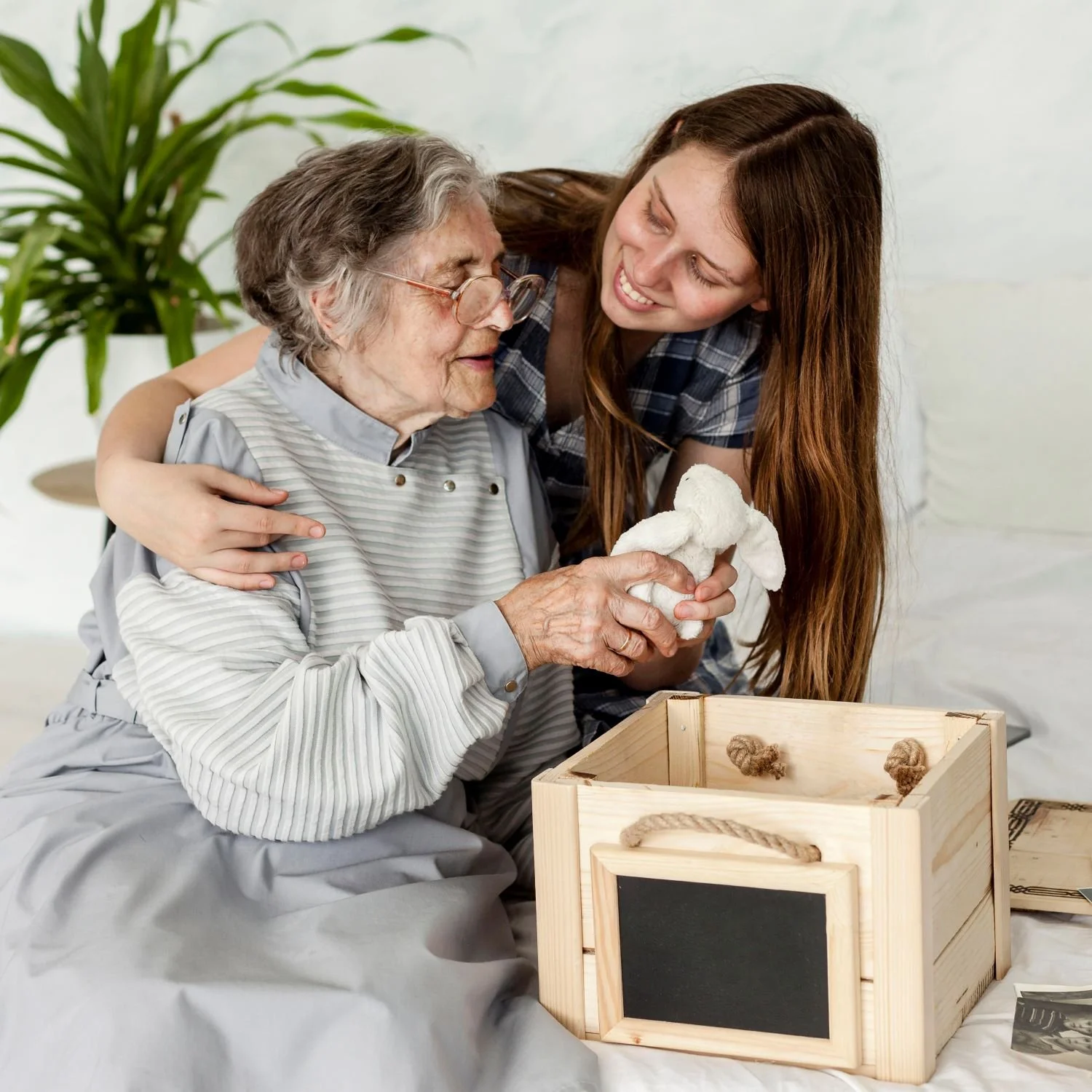 A young woman hugging an elderly woman and handing her a stuffed animal, with a wooden box on the table nearby.