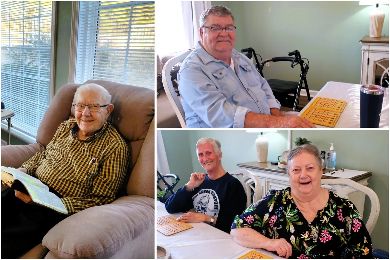 Group of four elderly people, two men and two women, sitting and smiling indoors around a table and a recliner chair, with one woman reading a book and a man using a walker in the background.