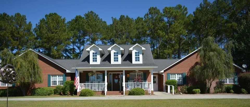 A large brick house with a grey roof and three dormer windows, a front porch with white railings, surrounded by green bushes and trees, with a clear blue sky in the background.