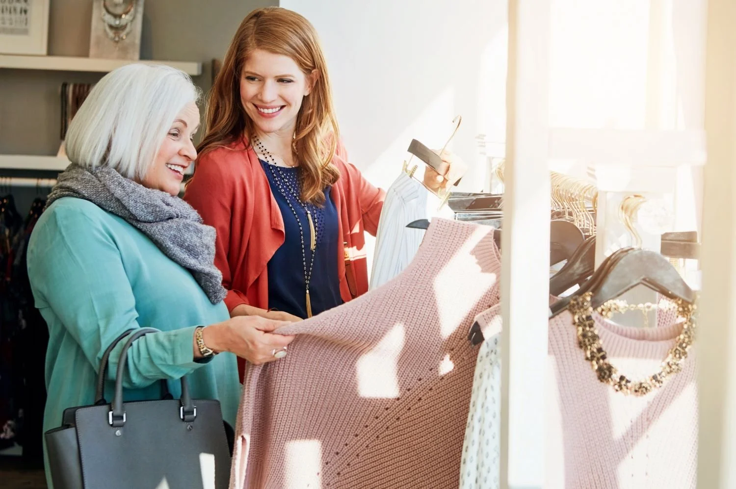 Two women shopping for clothes in a store, looking at a pink sweater with a necklace on a hanger.