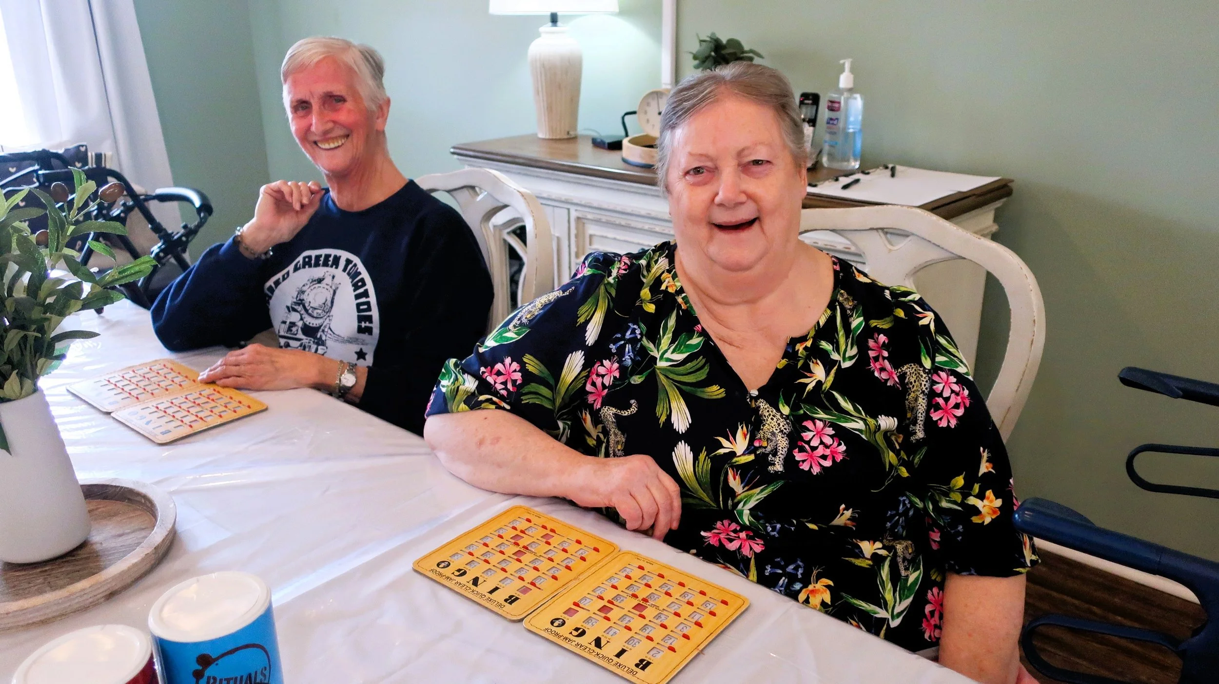 Two elderly women sitting at a table playing bingo, smiling, with bingo cards and drinks in front of them.
