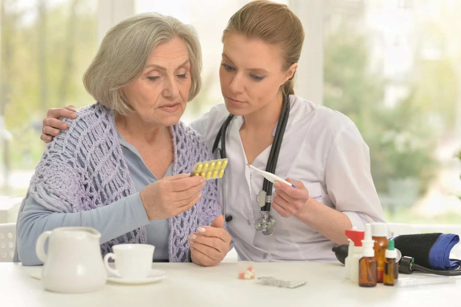 A young female healthcare worker, wearing a stethoscope around her neck, helps an elderly woman with medication at a table, with various medicines and a thermometer on the table.