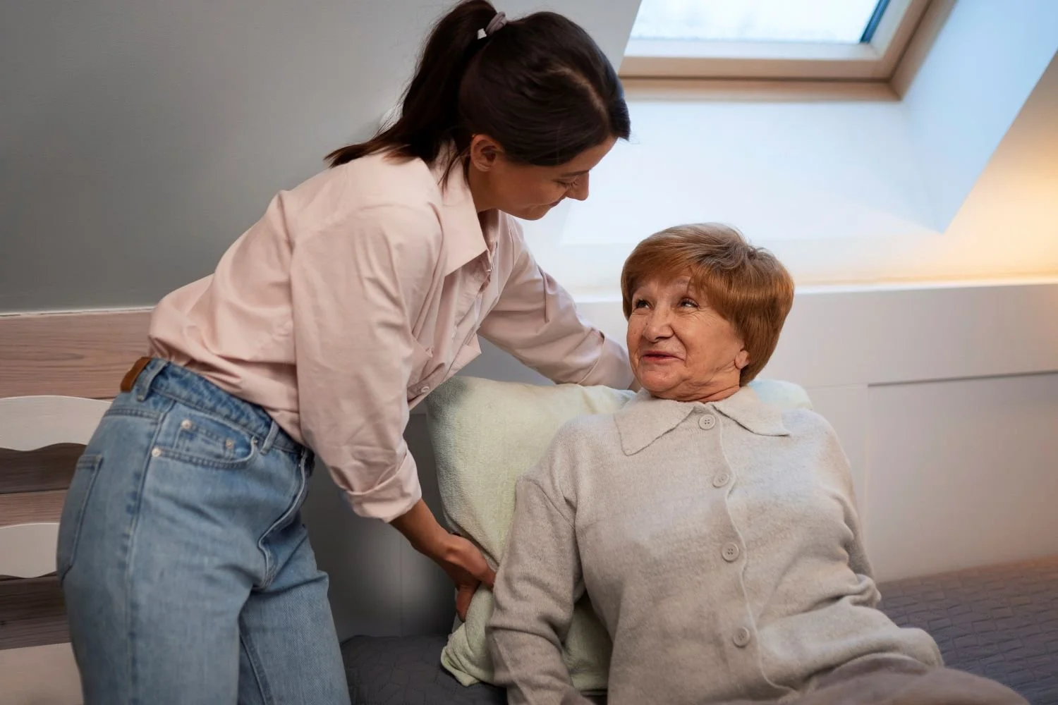 A young woman helps an elderly woman sit up in bed, smiling and engaging in conversation.