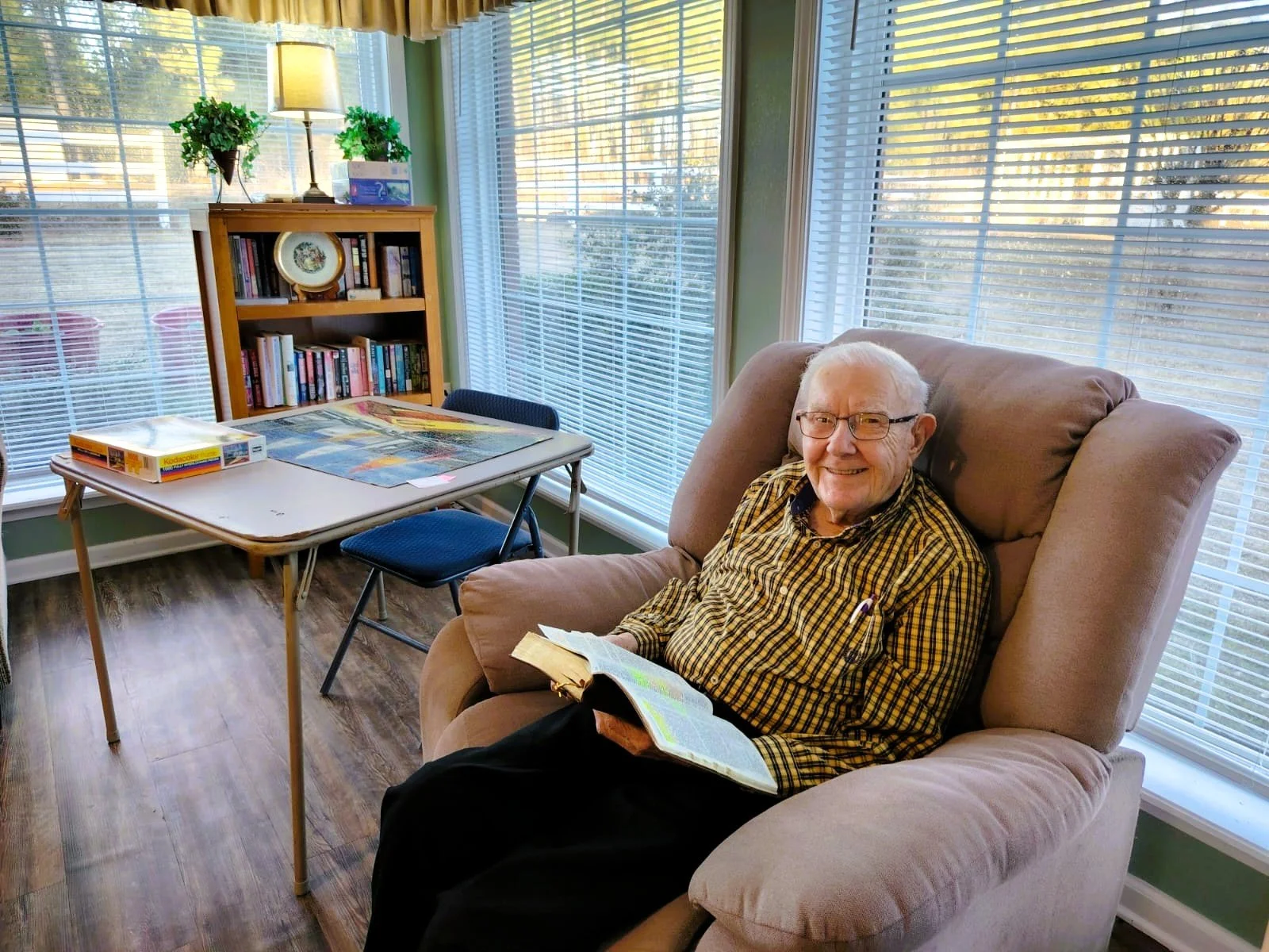 An elderly man with glasses smiling while sitting in a plush beige armchair and reading a book.