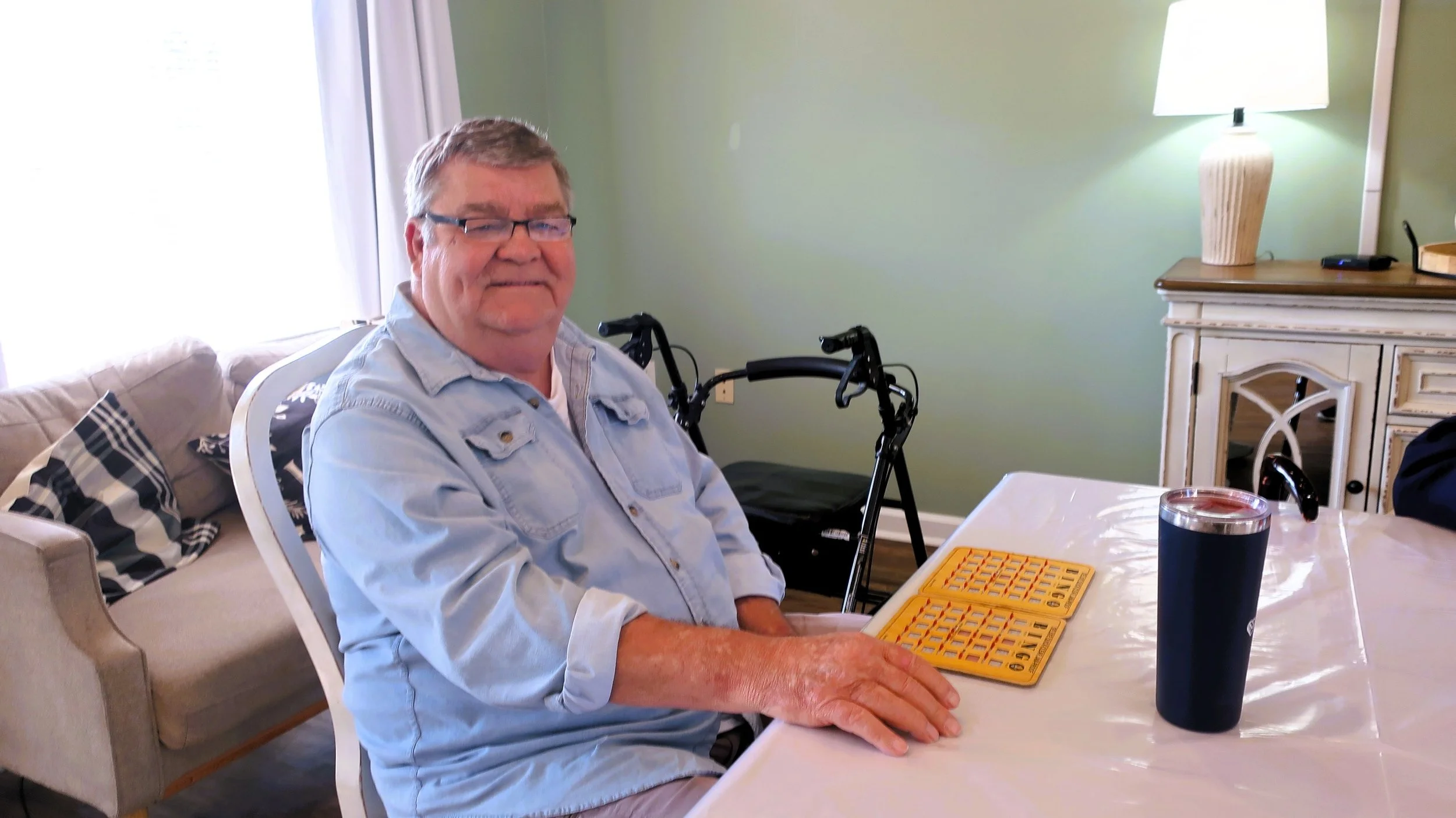 An elderly man with glasses and a light blue denim shirt sitting at a dining table with a yellow bingo card, a black tumbler, and a black walker behind him. A beige sofa with checkered pillows is in the background.