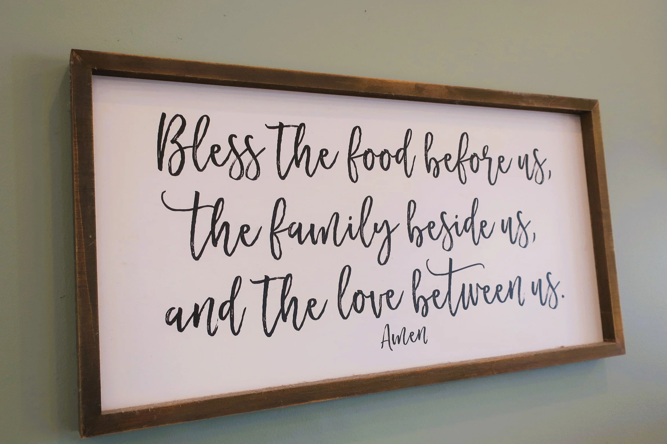 A wooden framed sign on a wall with a white background and black cursive text that reads: "Bless the food before us, the family beside us, and the love between us. Amen".