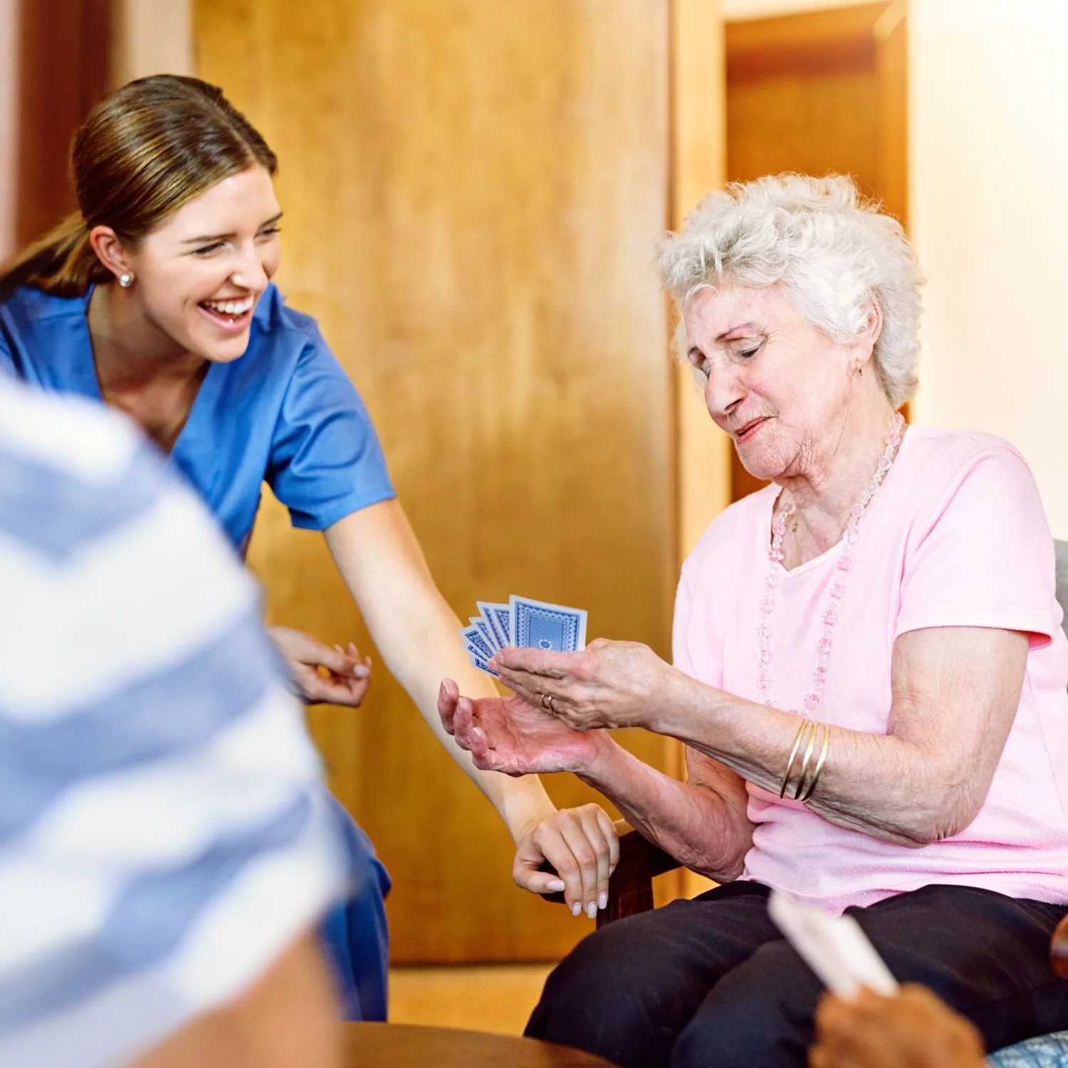 Young female nurse handing playing cards to an elderly woman sitting in a chair, in a warm and friendly setting.