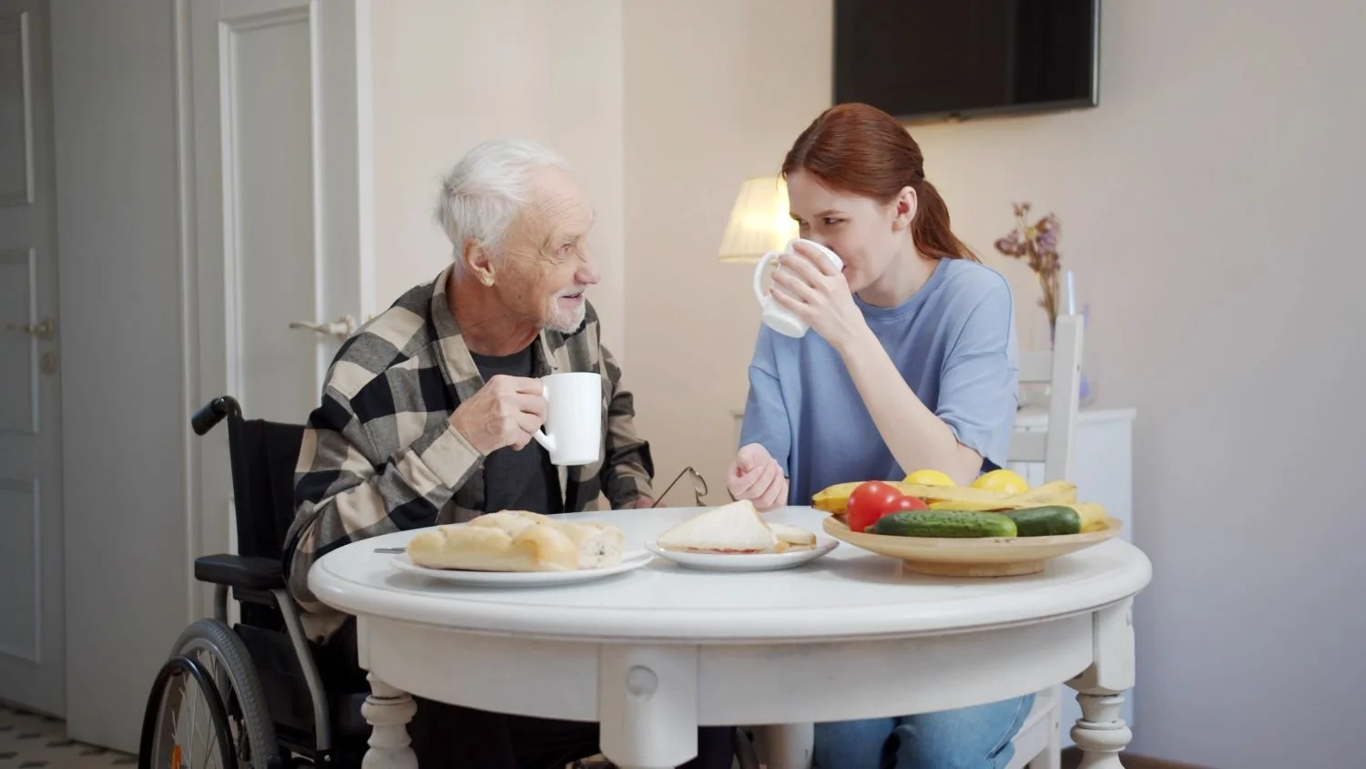 A young woman and an elderly man in a wheelchair share breakfast at a kitchen table, with the woman drinking from a mug and the elderly man holding a mug, smiling.