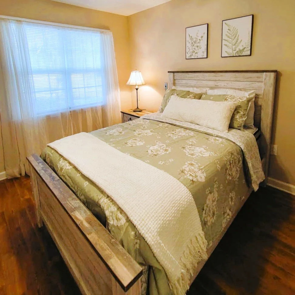 A bedroom with a wooden bed frame, floral bedspread, pillows, a bedside table, lamp, and framed botanical prints on the wall, illuminated by natural light from a window with sheer curtains.