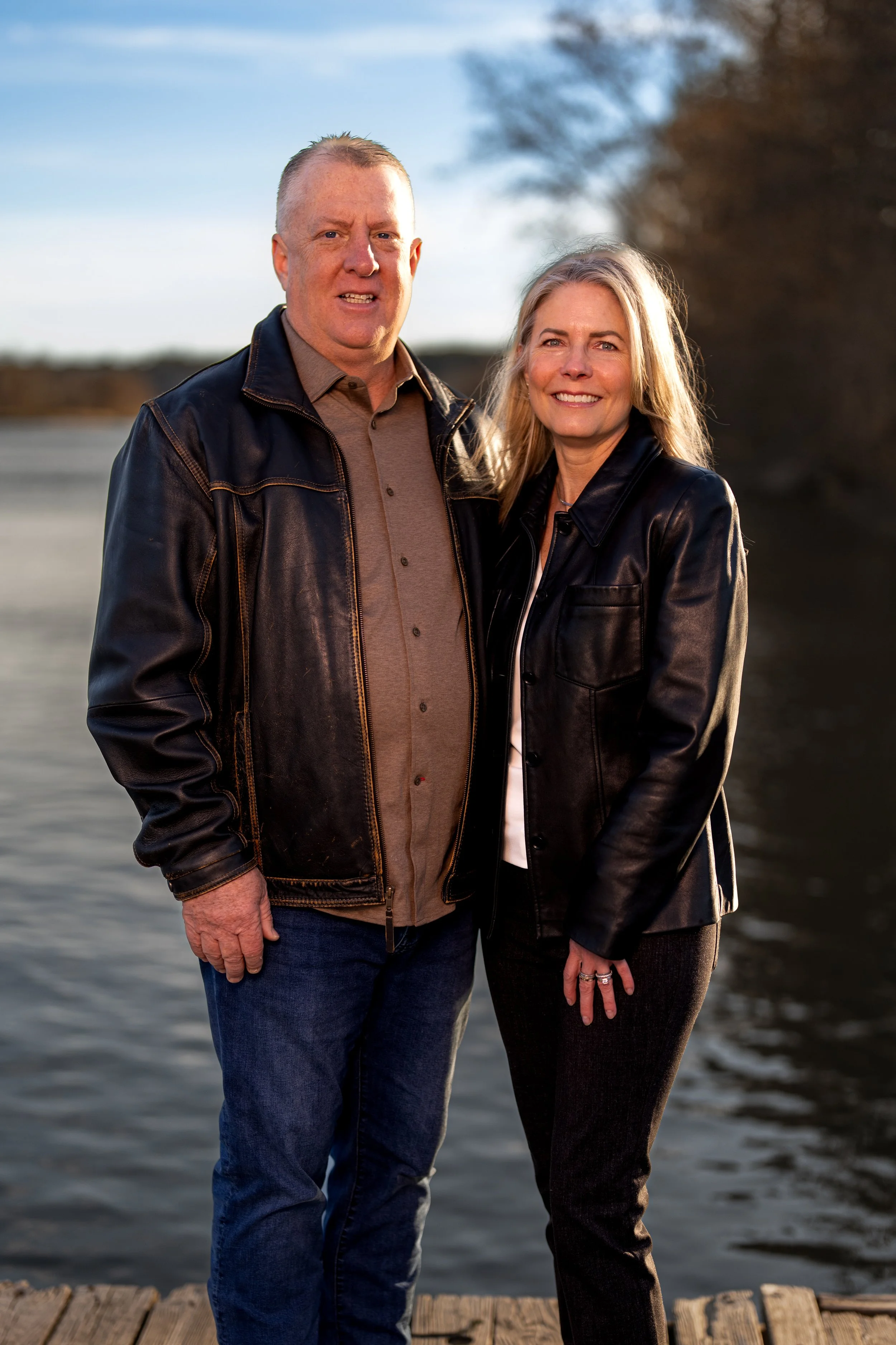 Portrait of a couple standing outdoors by a body of water. Couple positioned in the center of the frame, smiling broadly with a relaxed, genuine expression. Taken at Commons Ford Ranch Metropolitan Park in Austin by an Austin Texas photographer.