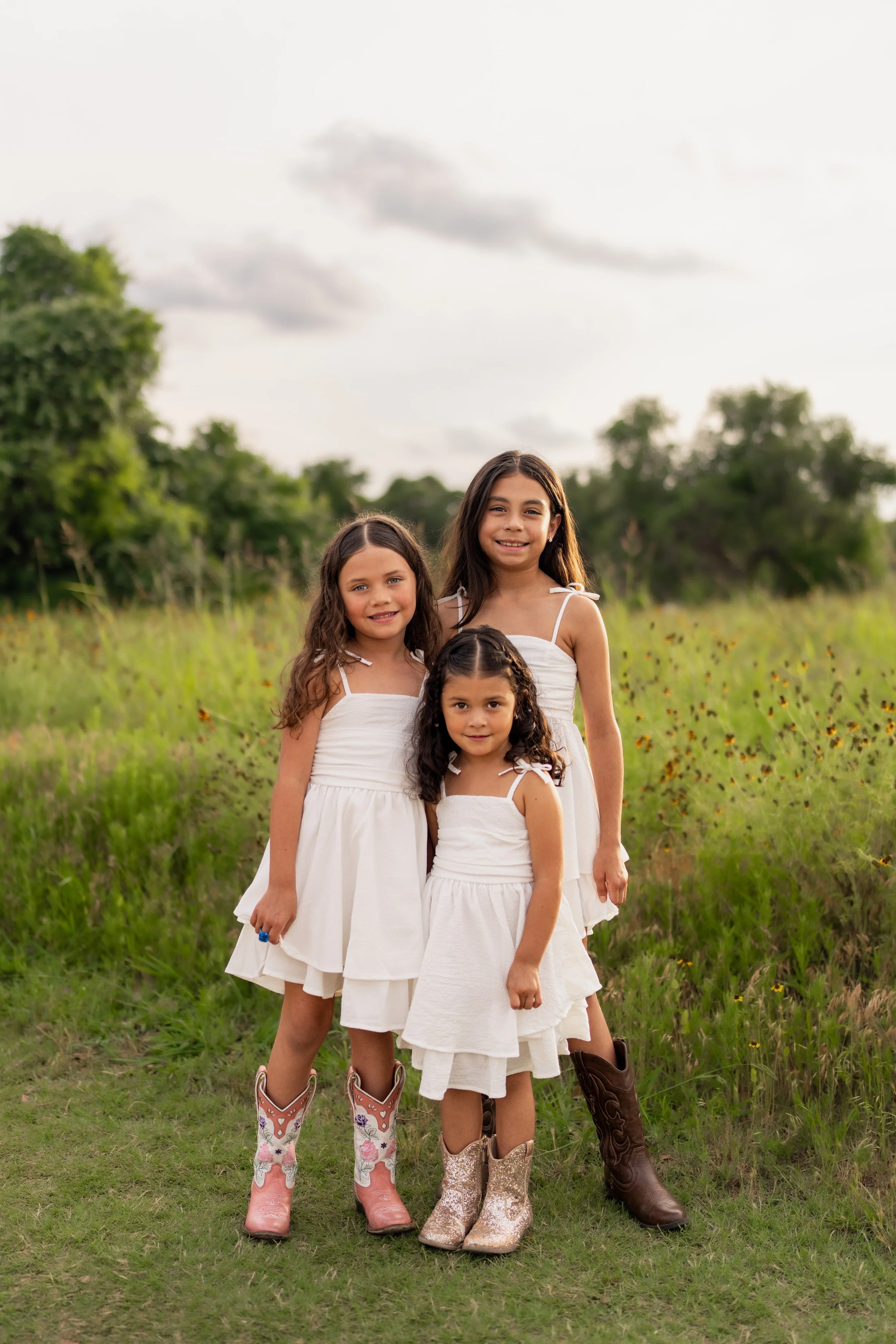 Three young girls standing outdoors in a field of tall grass and wildflowers, wearing white dresses and cowboy boots, smiling at the camera with trees and a cloudy sky in the background. Taken at Brushy Creek Lake Park Cedar Park Texas