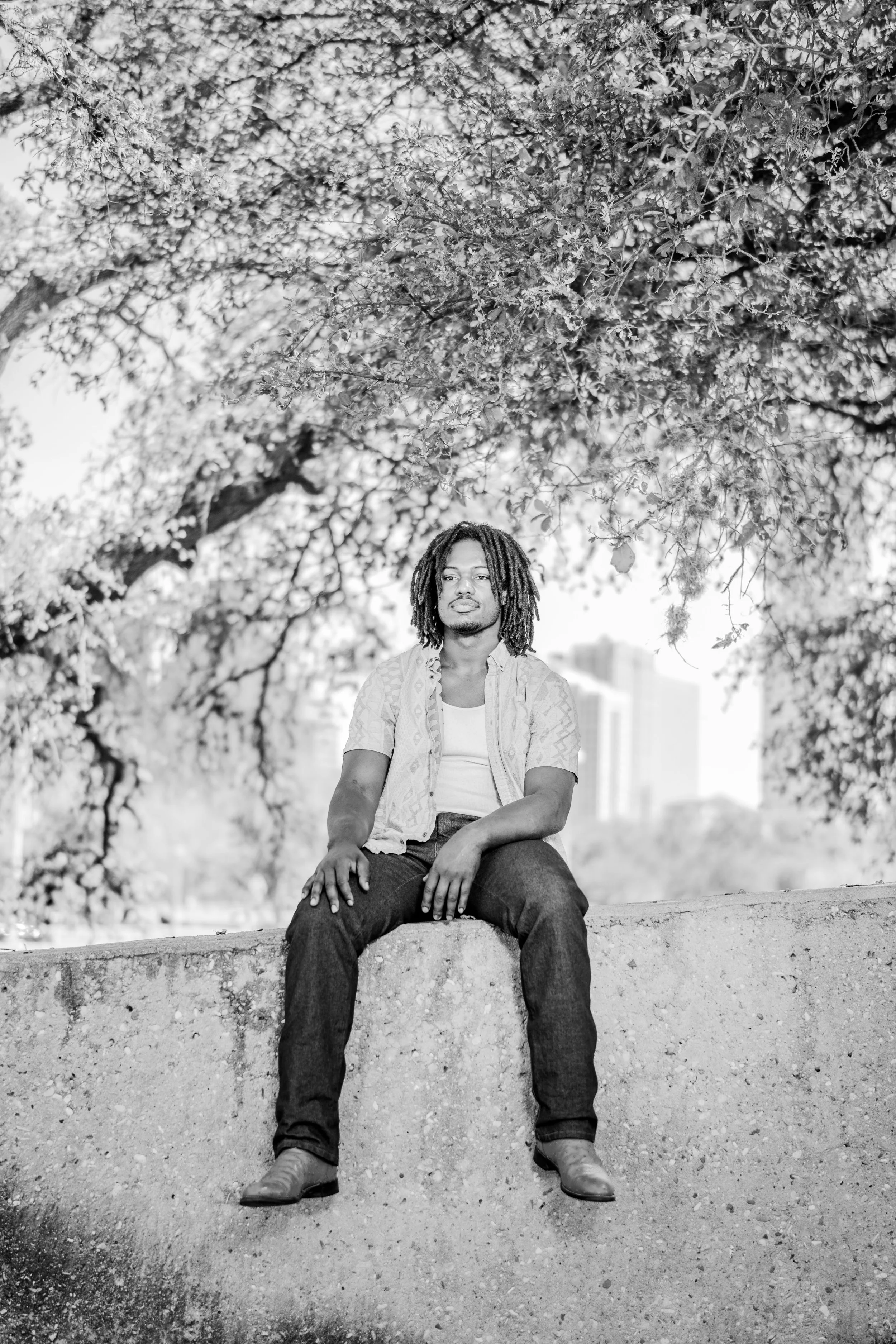 A man with dreadlocks in a western look sitting on a concrete wall while he looks off into the distance on a sunny day. Photo taken in Austin Texas by an Austin photographer.
