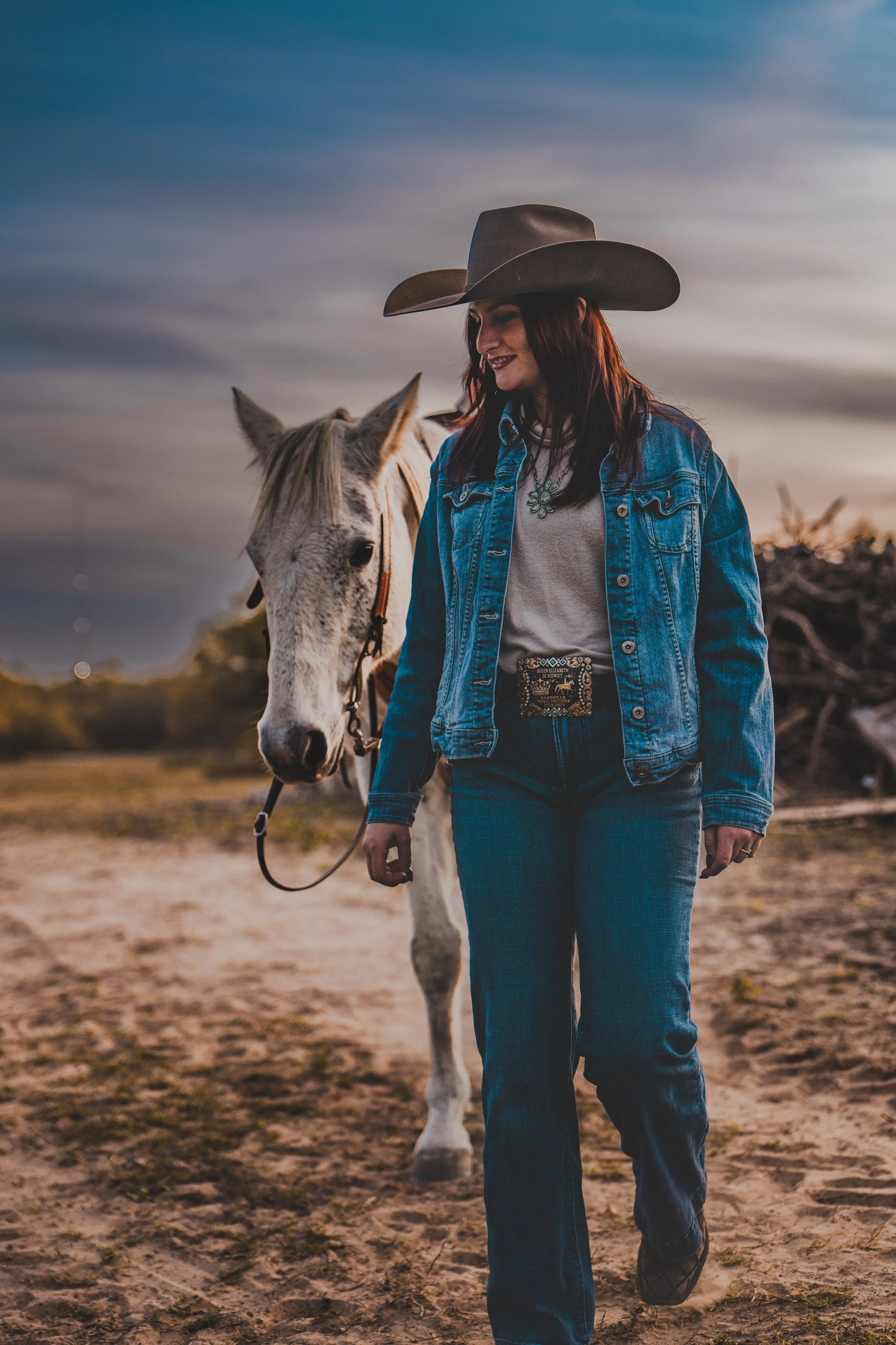 This photo captures a western-inspired portrait at golden hour. A woman wearing a wide-brimmed cowboy hat walks along a sandy path while leading a light-colored horse by the reins. Taken near Austin Texas.