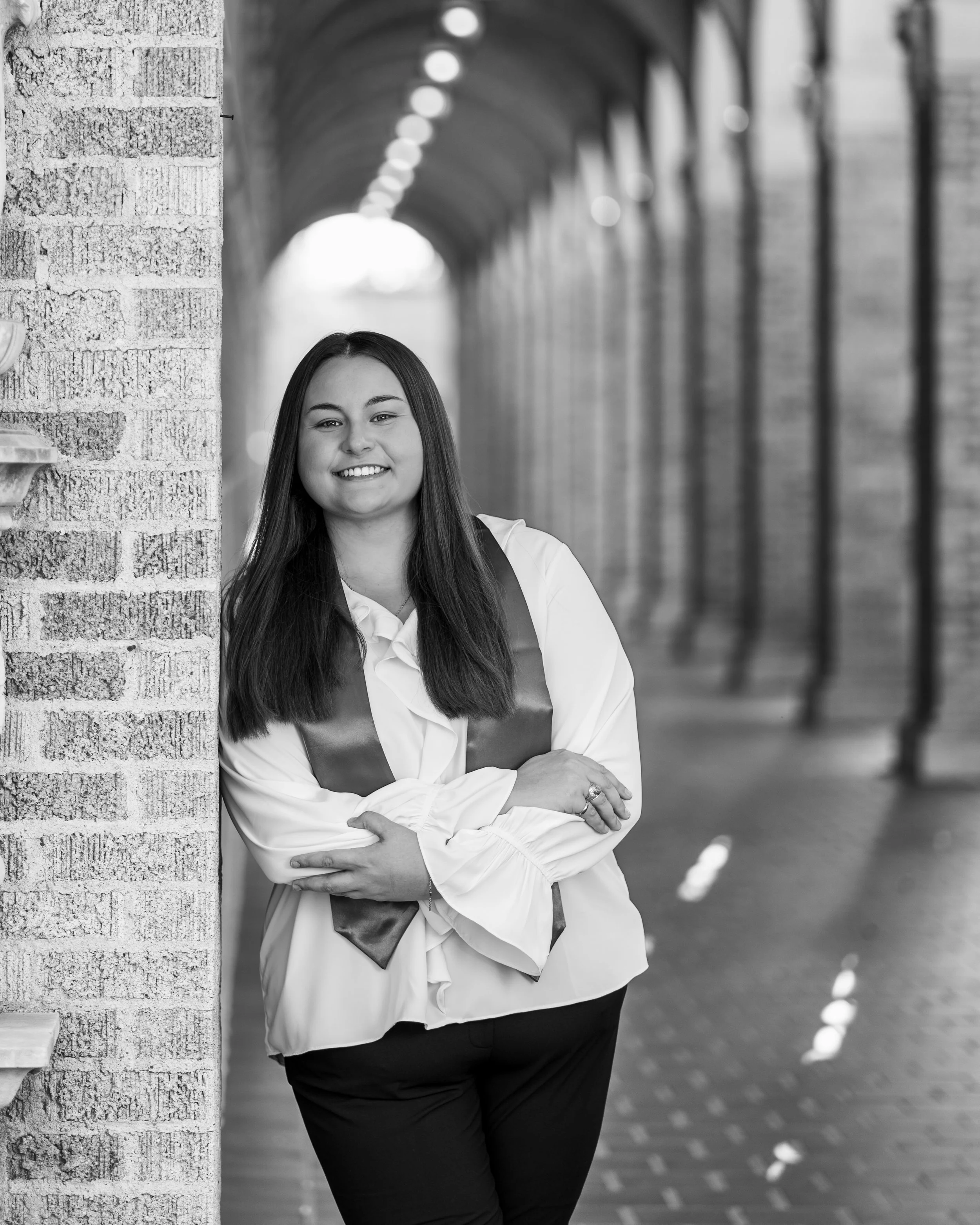 A young woman in business attire and tech stole stands outside in a covered walkway, smiling and leaning against a brick wall with leading lines and archway. Taken at Texas Tech University in Lubbock Texas