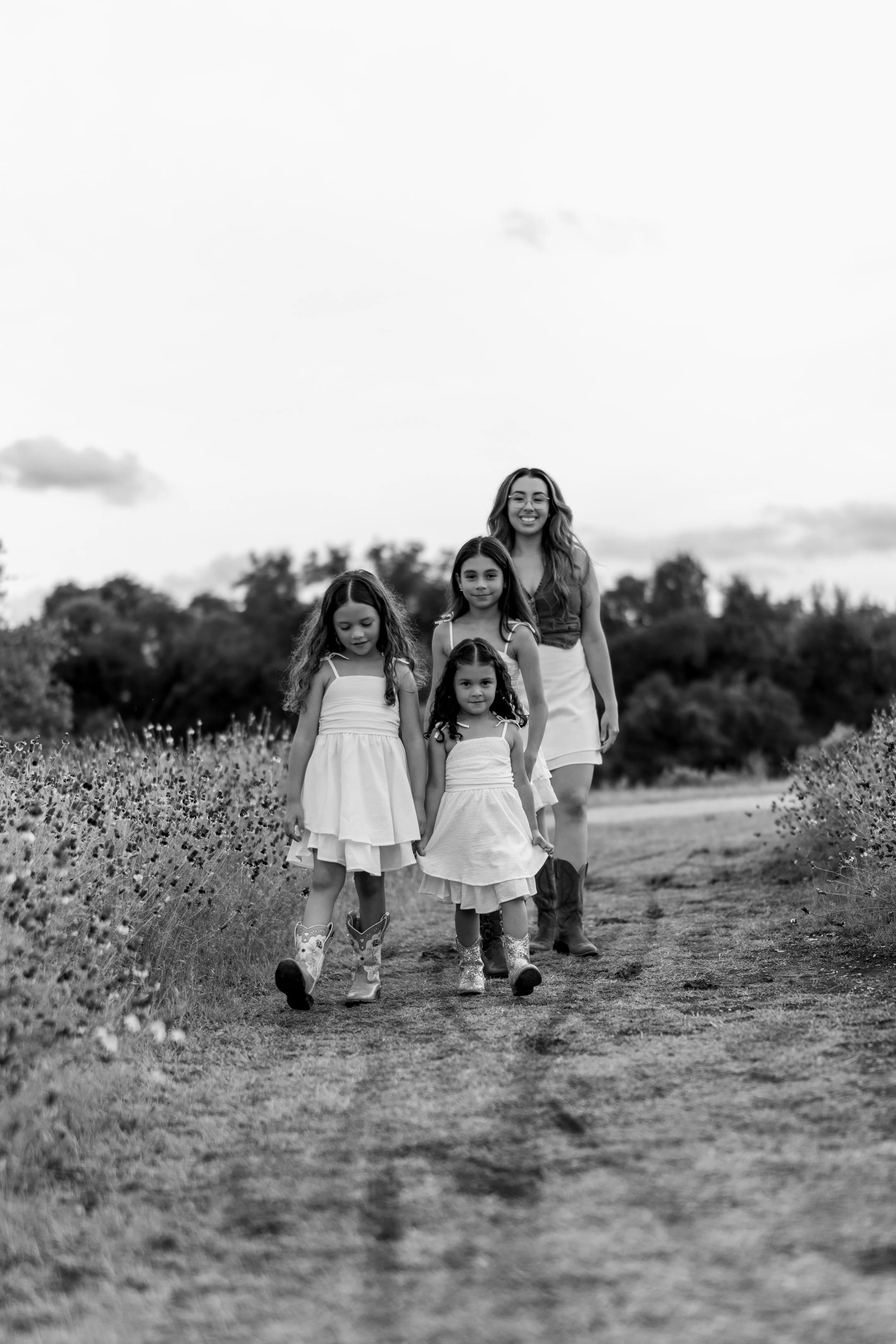 A woman and three young girls walking along a dirt path in a rural outdoor area, surrounded by grass and wildflowers, with trees and clouds in the background, all wearing dresses and boots in a black-and-white photo. Taken at Brushy Creek Lake Park.