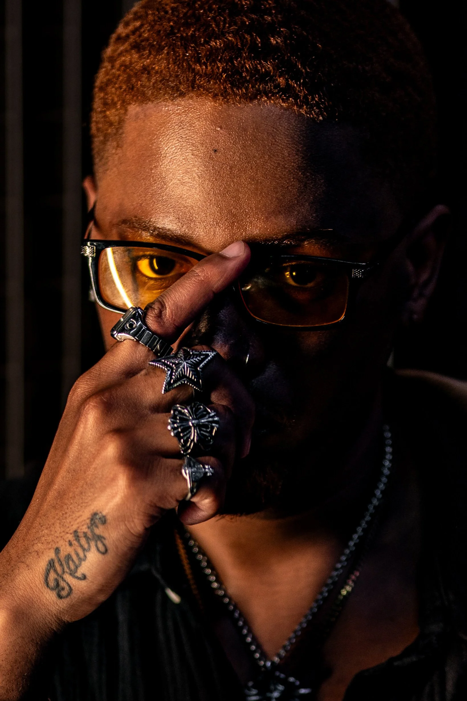 The subject is shown in a close-up shot, filling most of the frame. He’s adjusting a pair of amber-tinted glasses with his index finger placed at the bridge, partially covering one eye. Taken at High Five in Austin Texas.