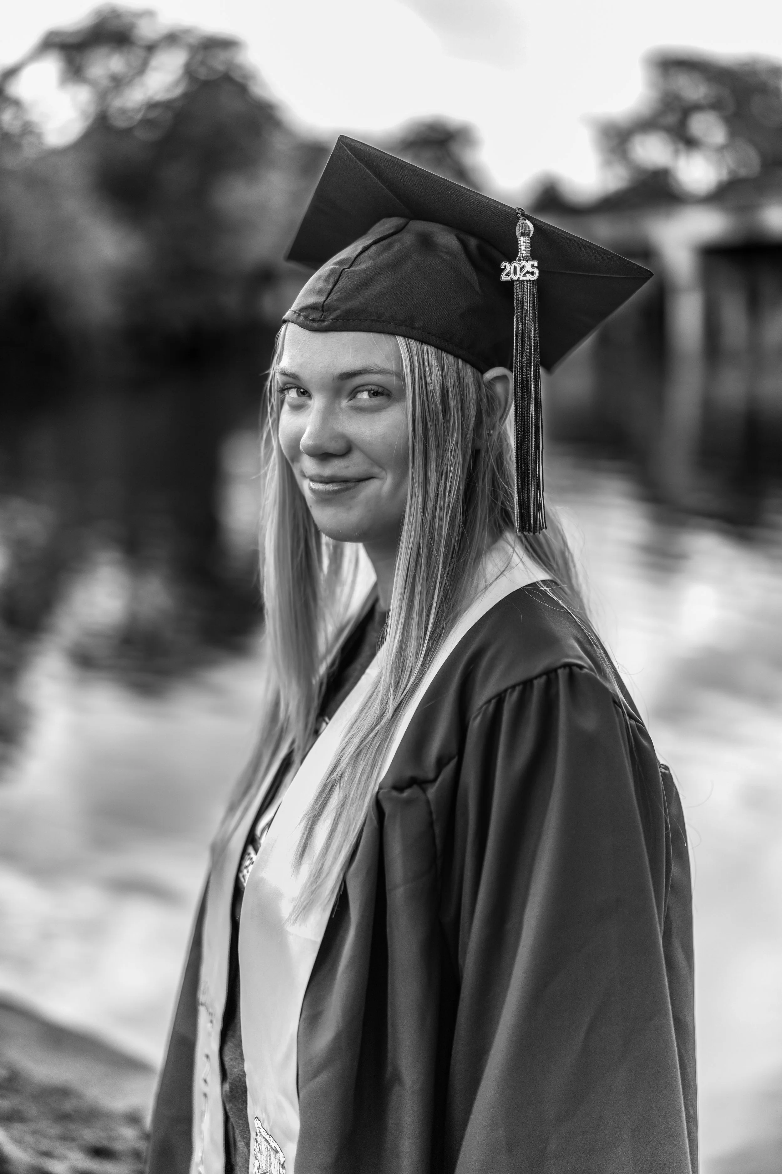 Black and white graduation photo of a young woman in a graduation cap and gown, standing outside near a body of water, smiling at the camera. Taken in San Marcos Texas by the San Marcos river.