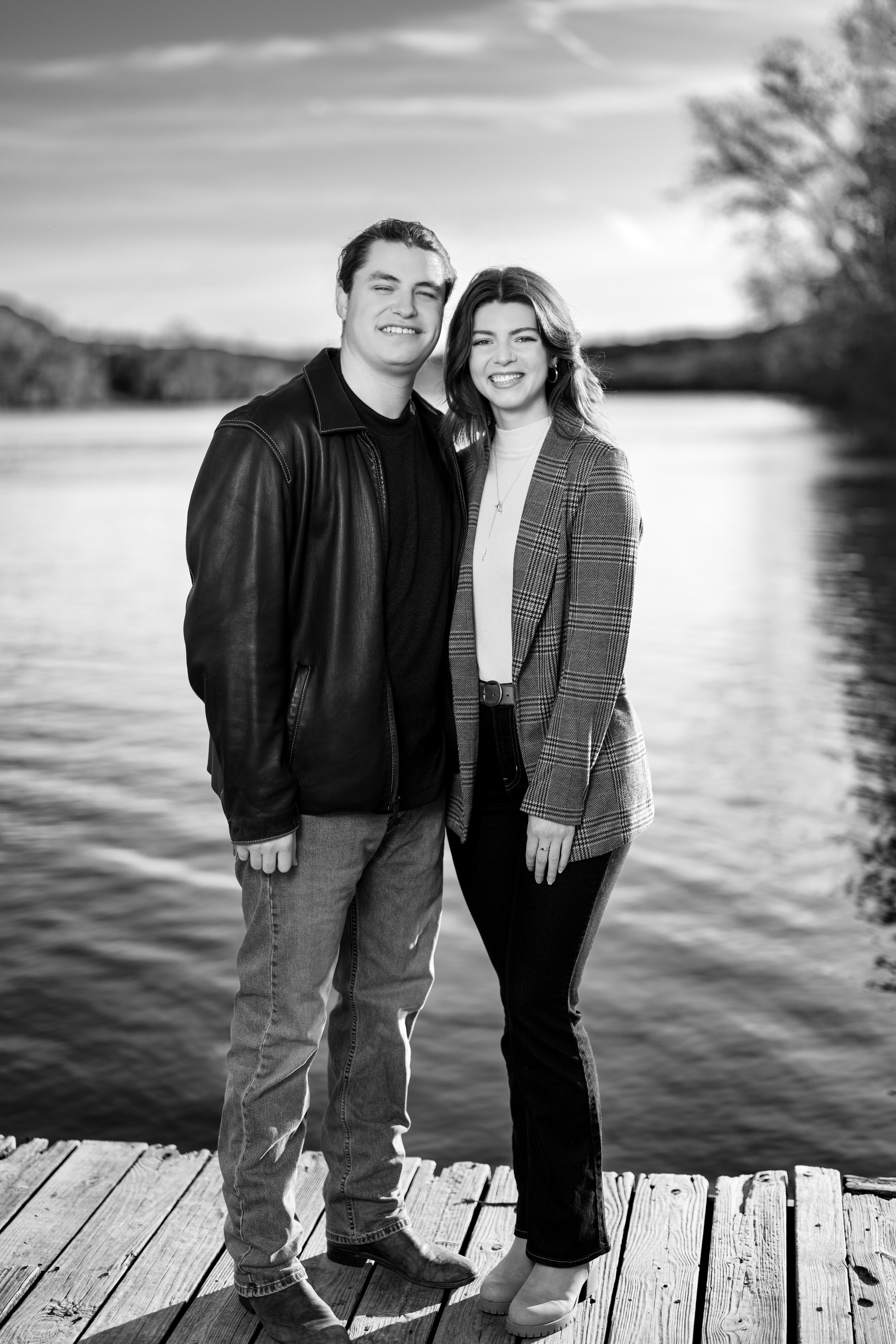 The image is a black-and-white portrait of a couple standing outdoors by a body of water. Couple positioned in the center of the frame, smiling broadly with a relaxed, genuine expression. Taken at Commons Ford Ranch Metropolitan Park in Austin Texas.