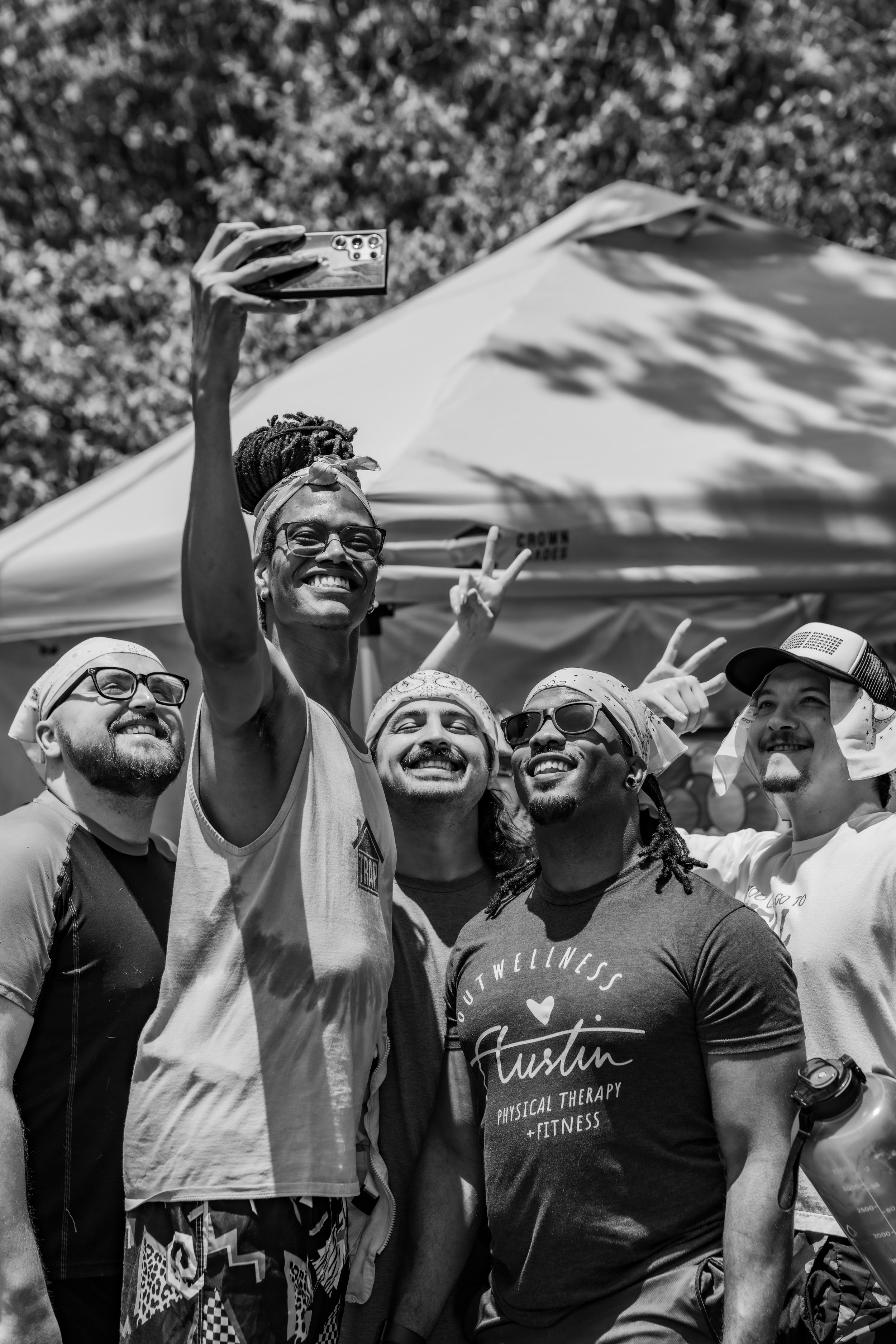A group of five diverse people smiling and taking a selfie outdoors under a canopy, some making peace signs, in black and white. Taken at a birthday party in Austin Texas with a field day theme.