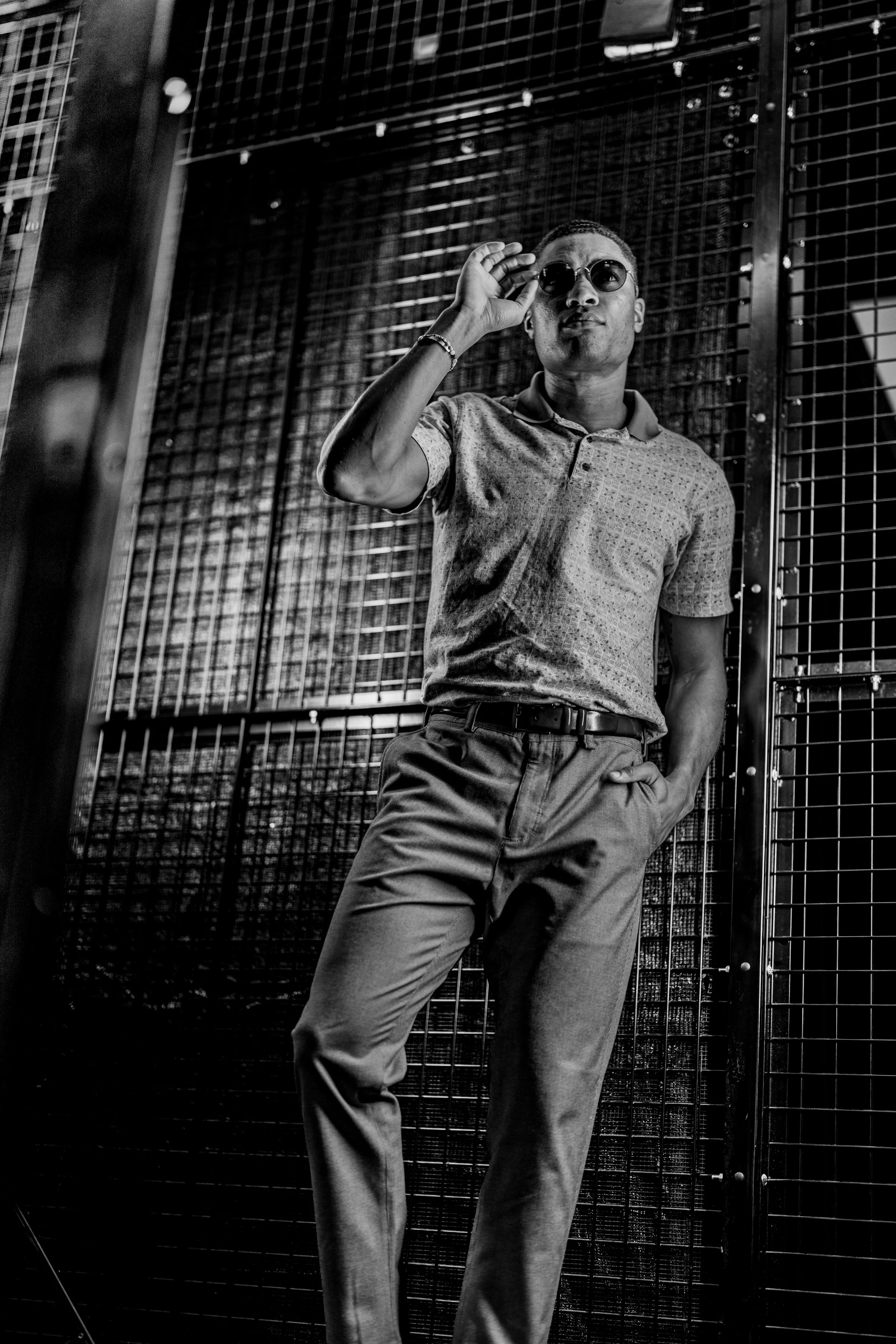 He’s standing in a relaxed but confident stance, one leg slightly bent with his foot resting against the wall. One hand is in his pocket, while the other is raised to adjust a pair of round sunglasses, adding a stylish, composed feel to the pose. Tak