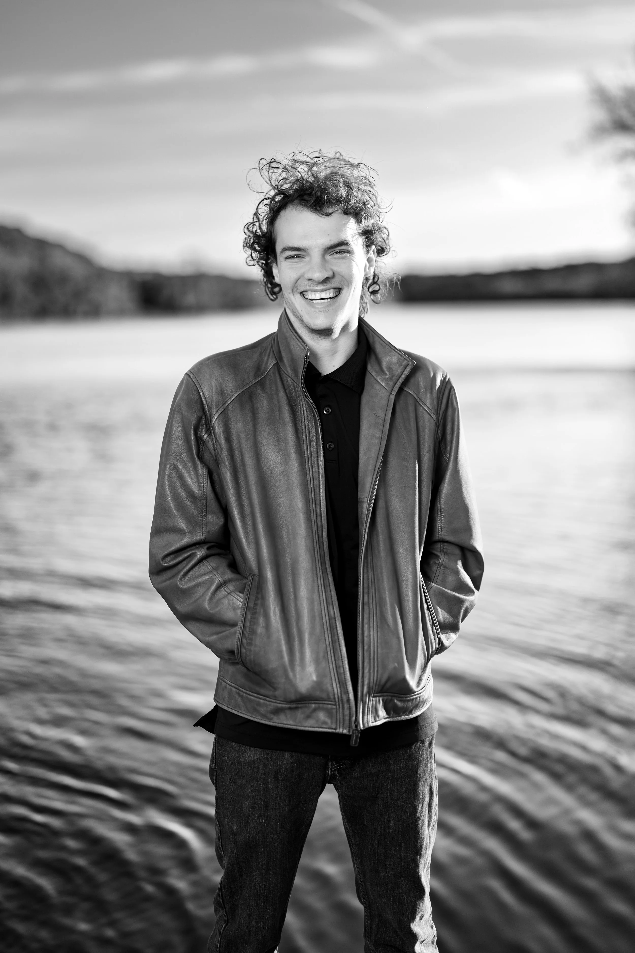 The image is a black-and-white portrait of a young man standing outdoors by a body of water. He’s positioned in the center of the frame, smiling broadly with a relaxed, genuine expression. Taken at Commons Ford Ranch Metropolitan Park in Austin Texas