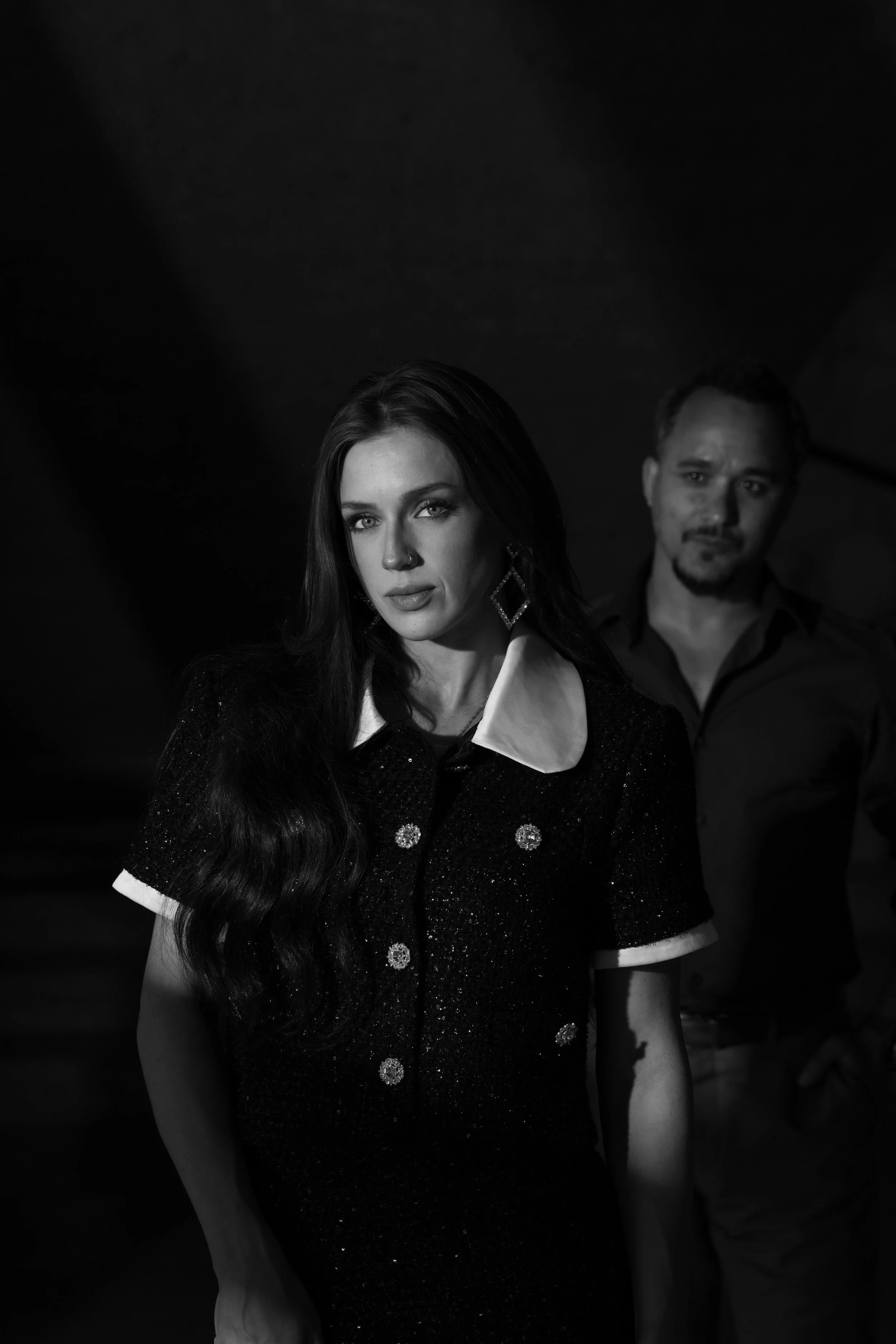 A black and white portrait of a woman with long dark hair, wearing earrings, a dark dress with a white collar, and standing in the foreground, with a man in the background. Taken in Austin Texas.
