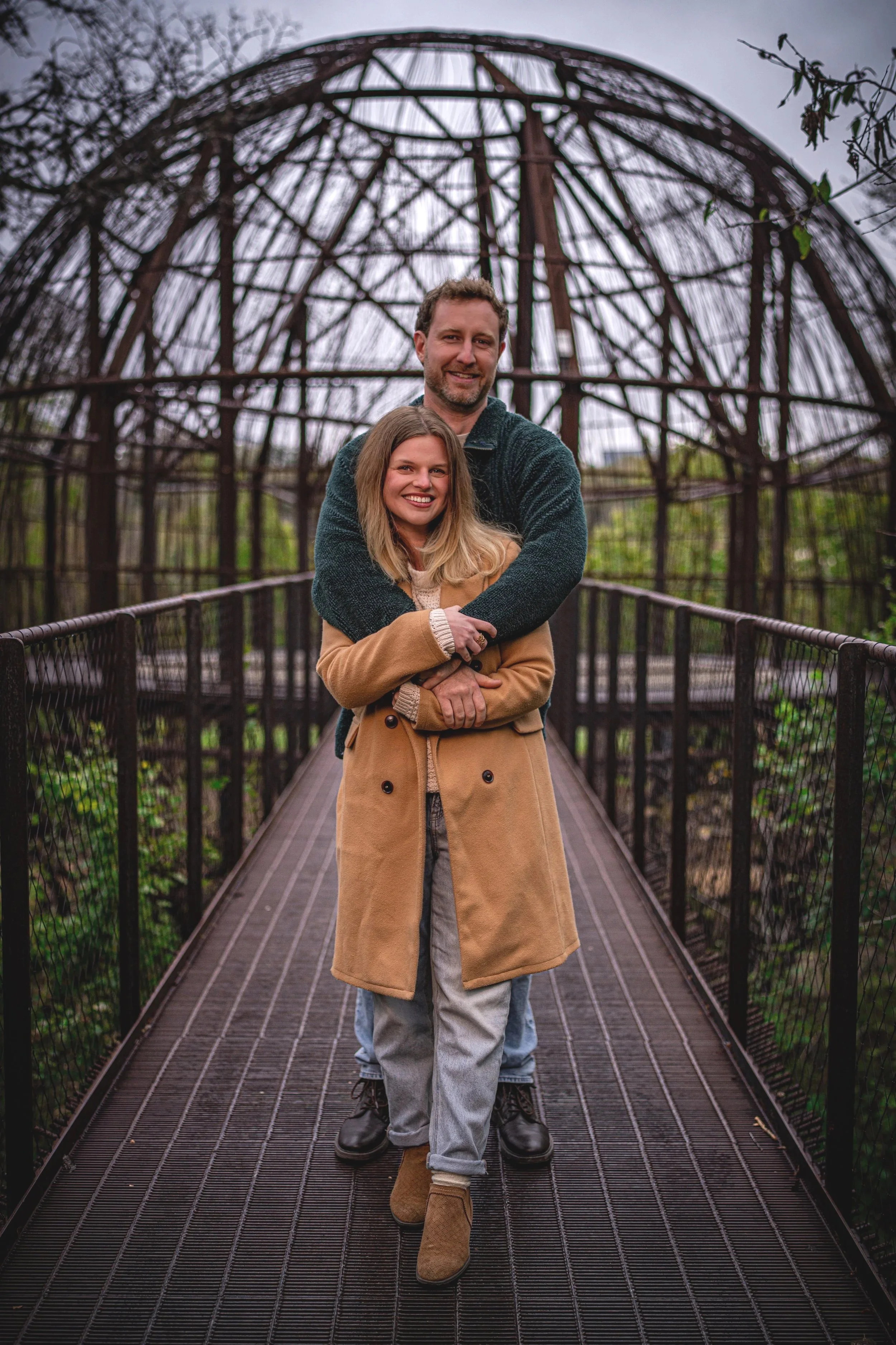 A smiling couple hugging on a bridge under a metal arched structure, outdoor setting. Photo taken at the treehouse in Austin Texas.