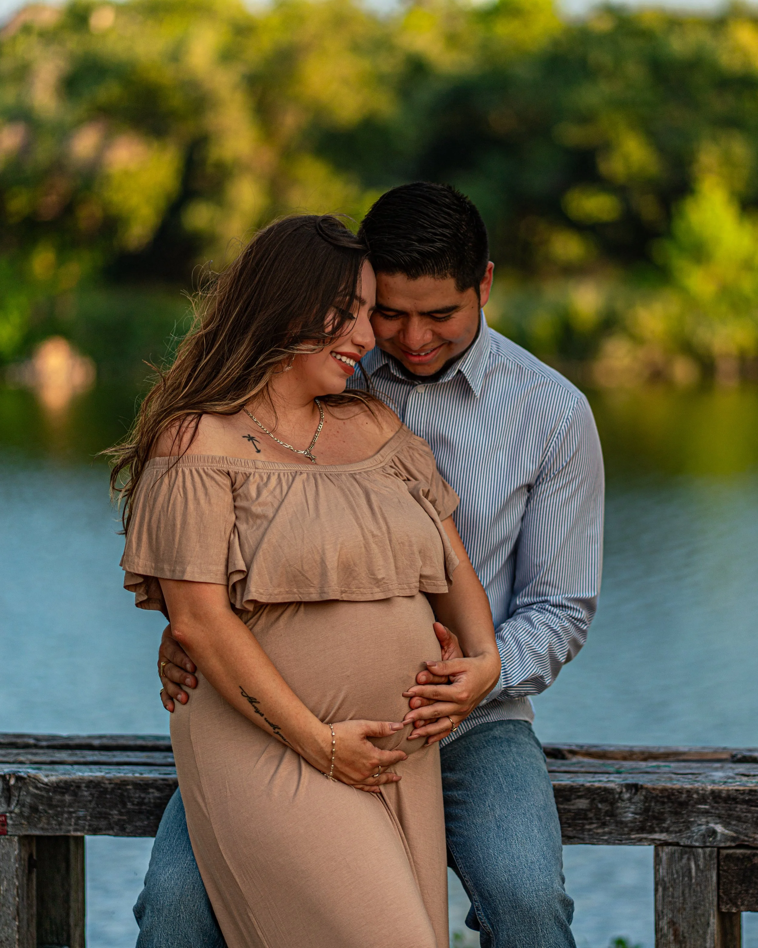 A beautiful pregnant woman and her husband sit on a bench in front of a lake. Their heads touch each others while the man tenderly touches the womans belly.