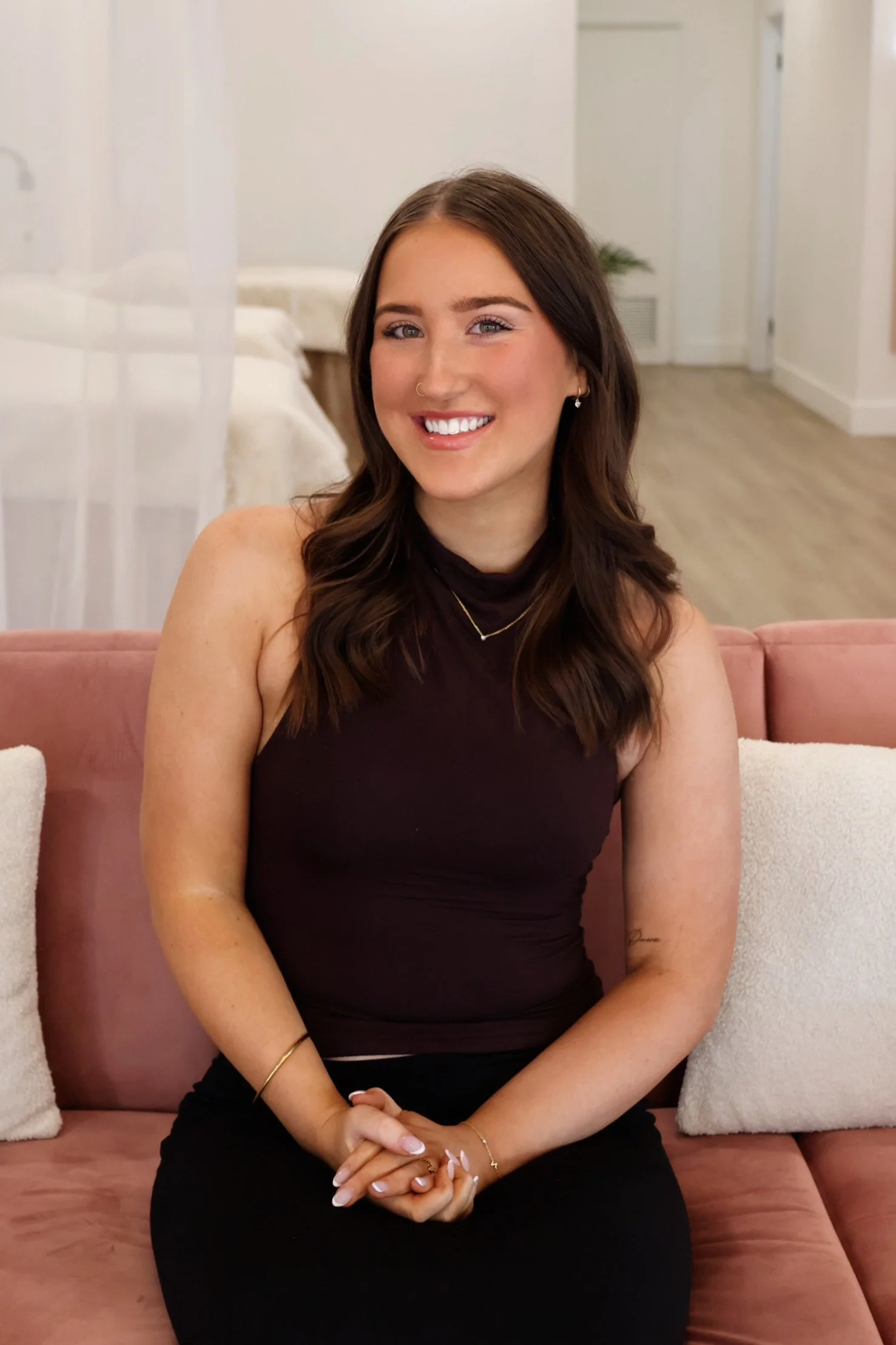 A smiling woman with long brown hair, wearing a sleeveless dark top, two necklaces, and earrings, sitting on a pink couch in a well-lit room with white walls and a wooden floor.