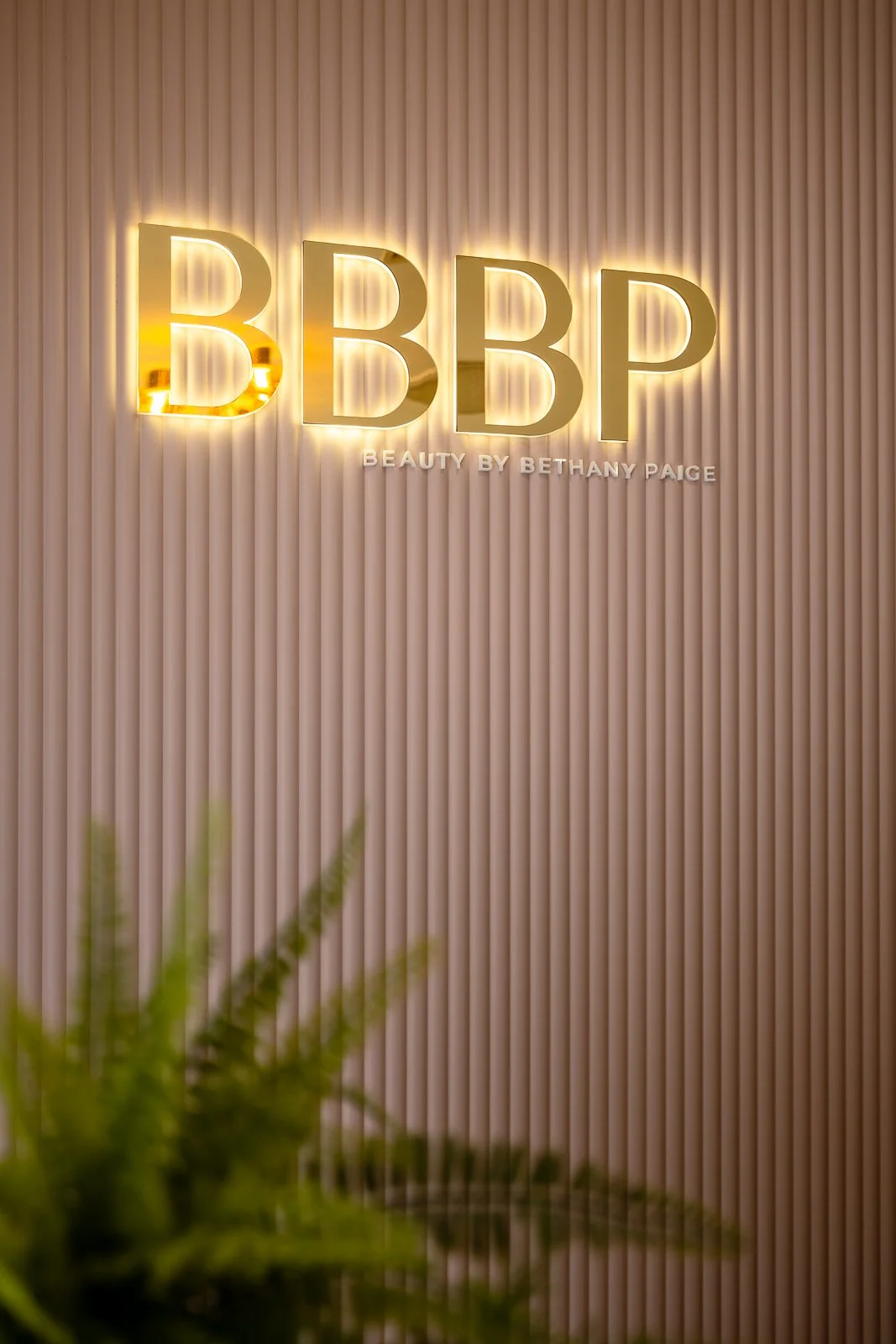 Sign for a beauty salon called B.B.P. illuminated with backlighting, mounted on a textured beige wall, with the phrase 'Beauty by Bethany Paige' underneath, and some greenery at the bottom edge.