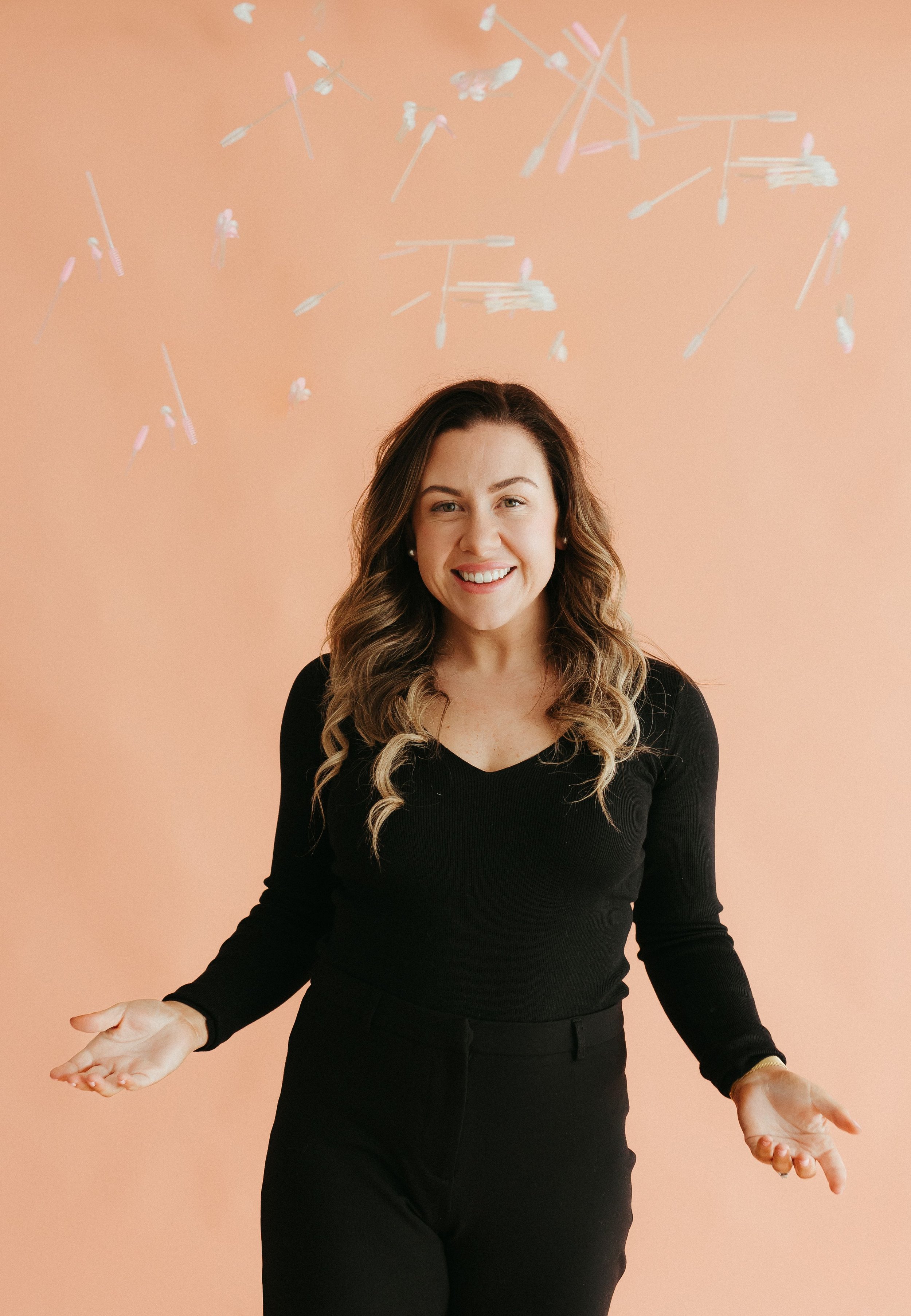 A woman with long, wavy brown hair, wearing a black long-sleeve top and black pants, standing against a peach-colored background with scattered makeup swabs floating above her head, smiling with her arms extended outward.