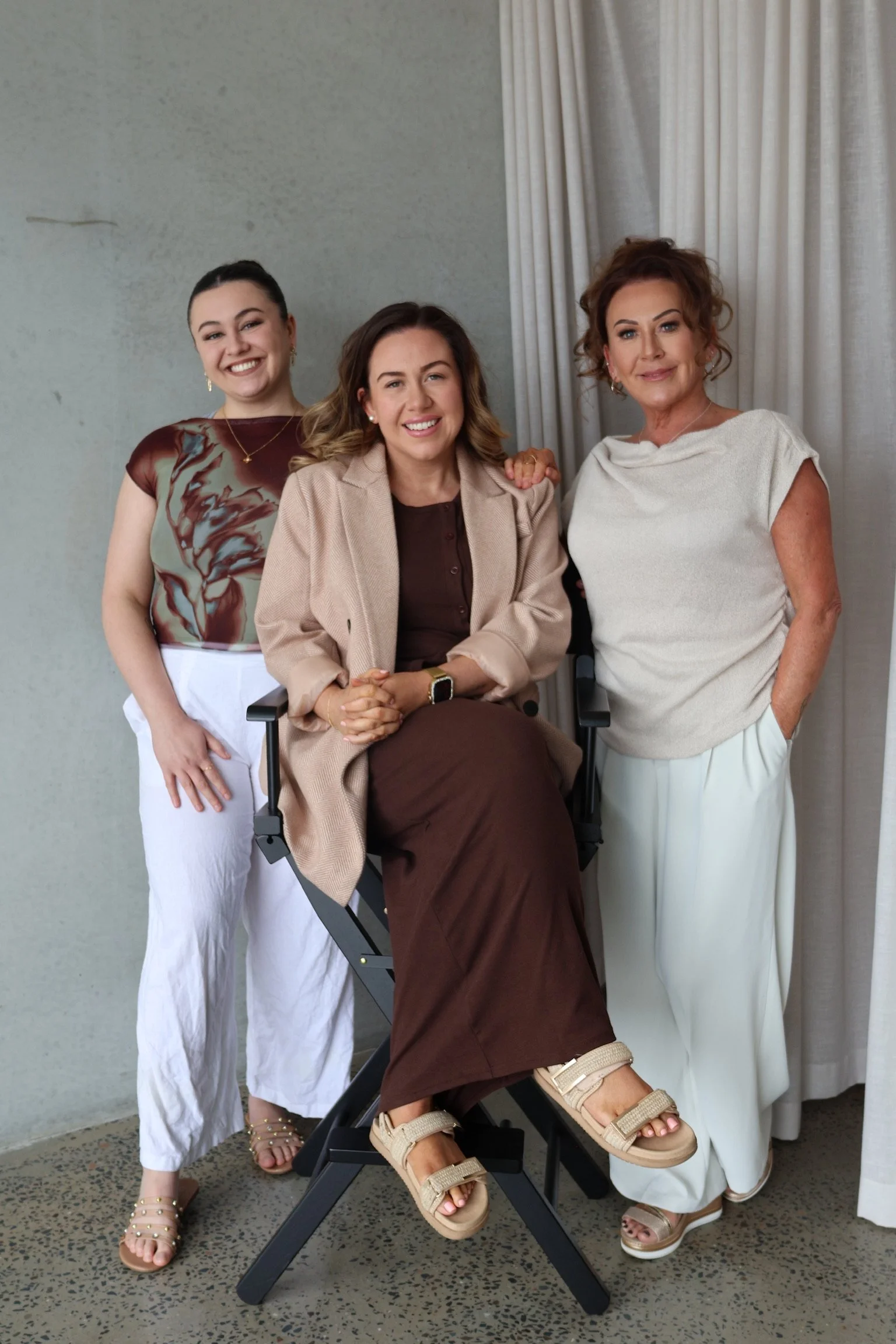 Three women standing around a seated woman in a wheelchair. The woman in the wheelchair has brown hair, wears a beige blazer over a brown dress, and beige sandals. The woman on the left has dark hair in a ponytail, wears a patterned top and white pants. The woman on the right has curly hair, wears a white top and white pants. They are indoors in front of a plain wall with a curtain.