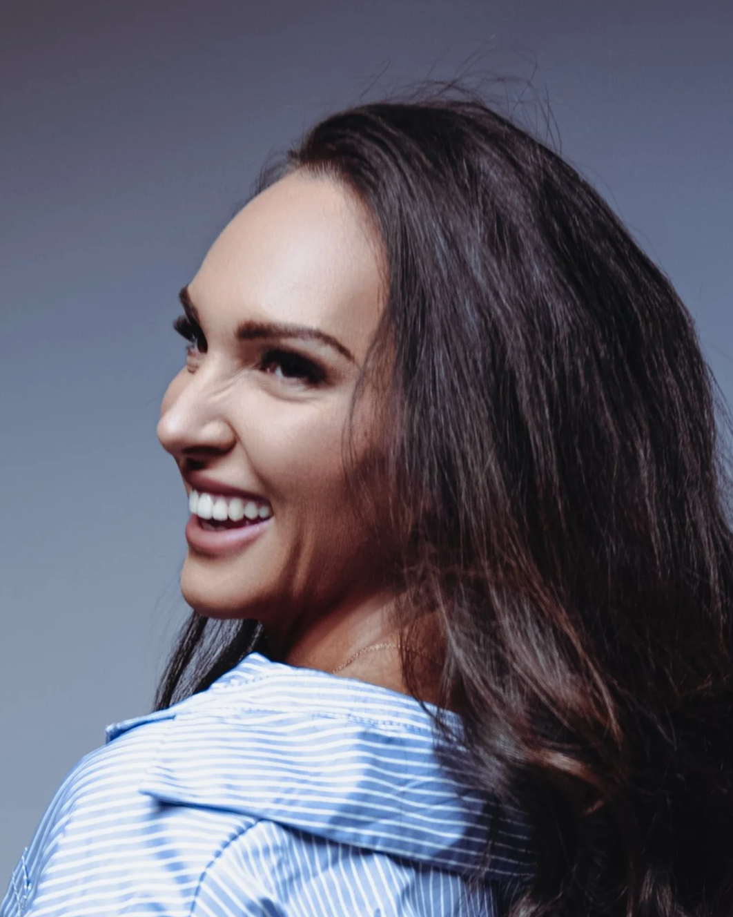 A woman with long, dark, wavy hair smiling and looking to the side, wearing a light blue striped shirt against a plain gray background.