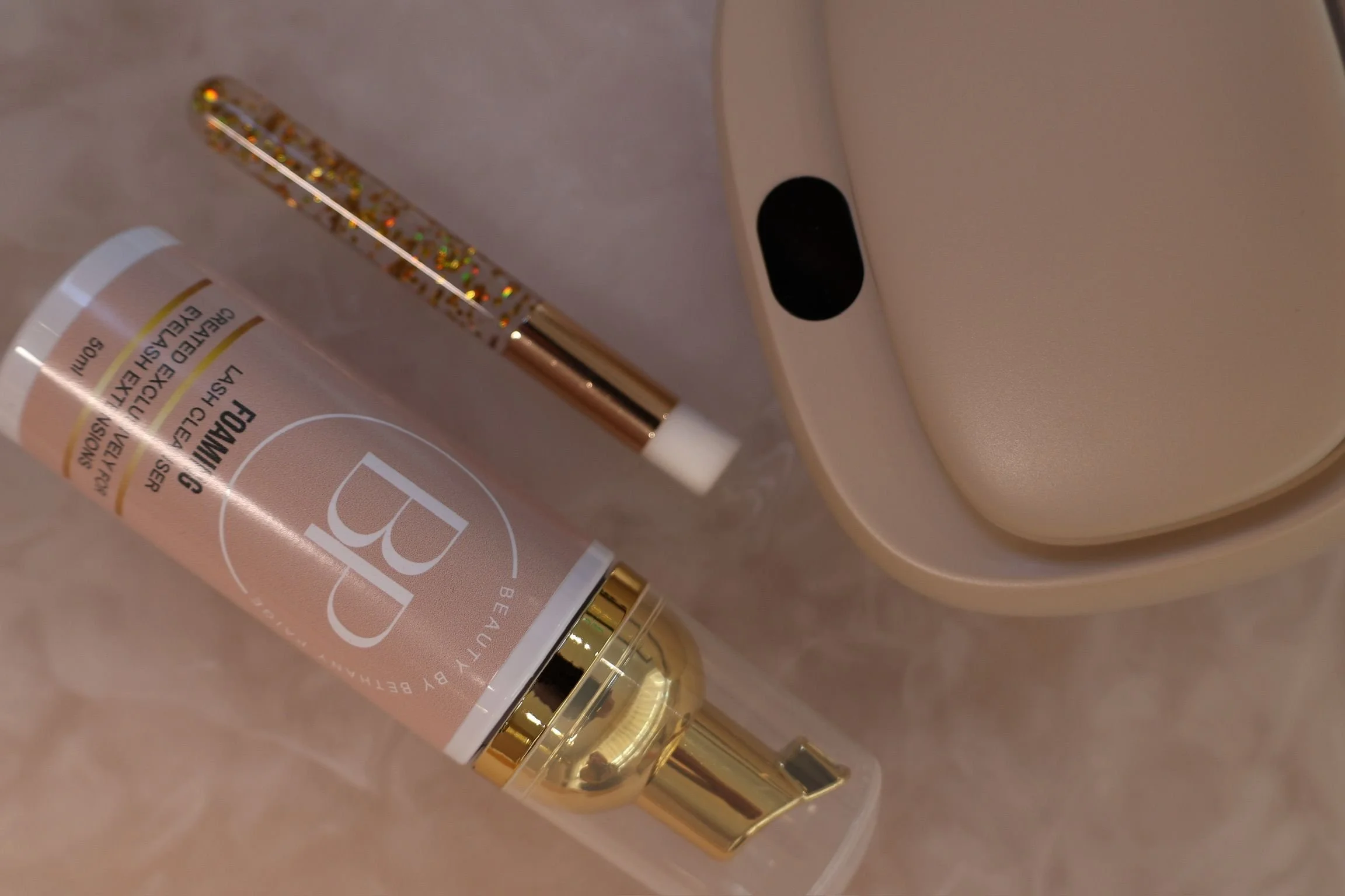 A tube of beige cream with white, black, and gold accents, placed on a soft pink surface. Part of a device with a black oval button and a white body is visible in the background.