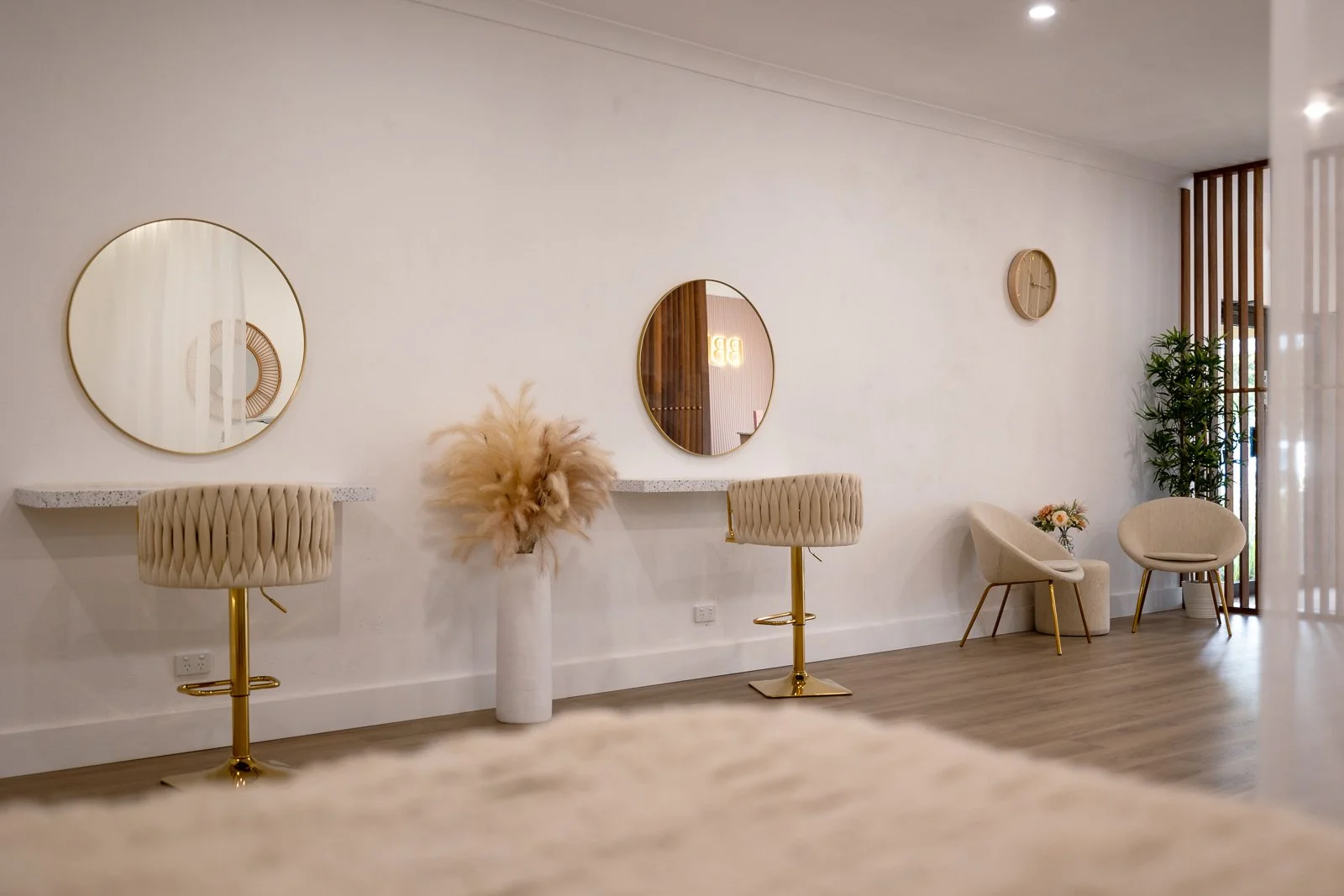 Modern interior space with cream chairs, round mirrors, a flower arrangement, and a tall green plant against a white wall.
