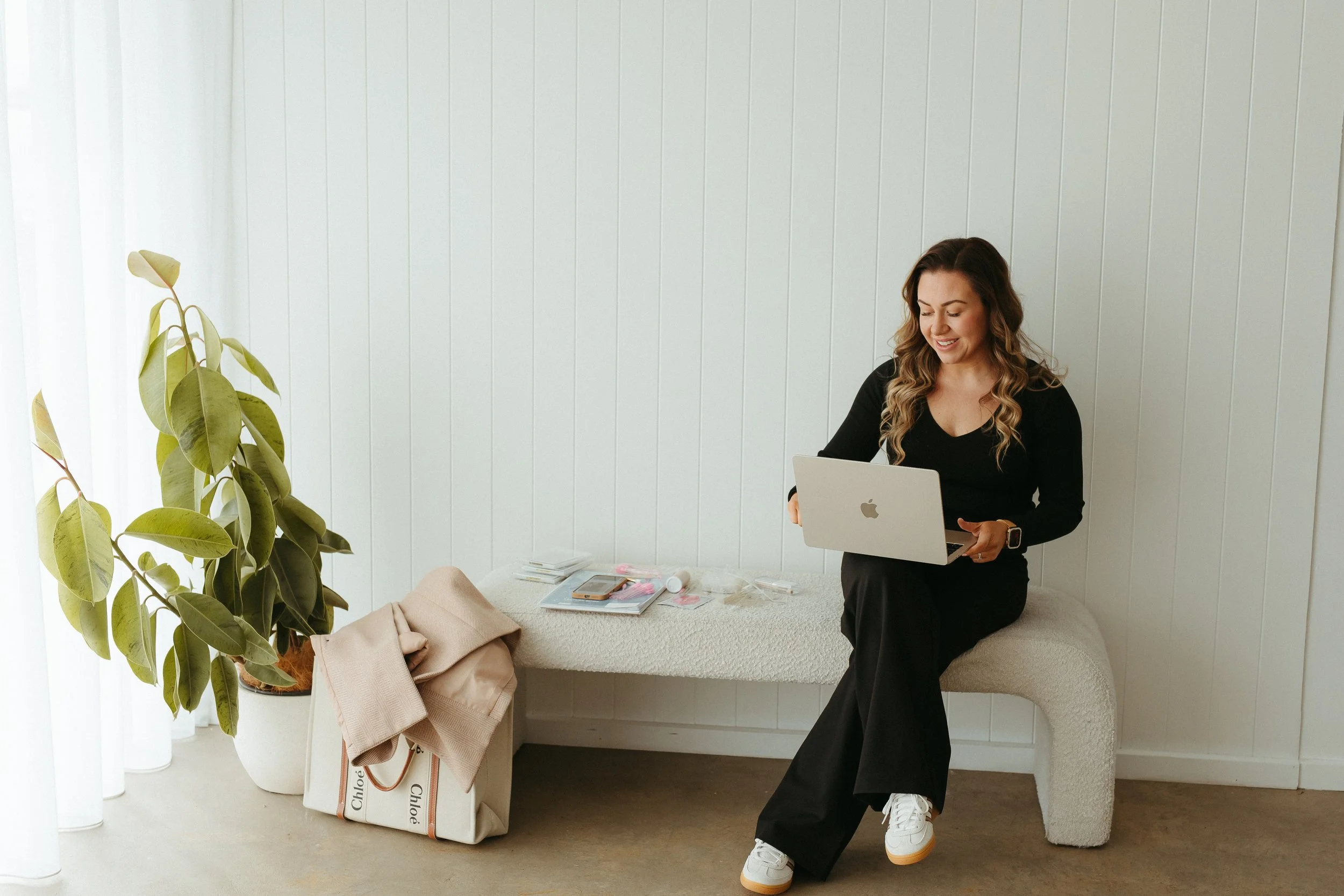 A woman with long wavy hair wearing a black top and black pants is standing in a bright room with white walls. She is smiling and holding a silver MacBook laptop. There is a large green houseplant on the left, and a beige bag with a beige coat and some shopping bags on a white cushioned bench. Items such as a smartphone, notebooks, and small containers are on the bench.