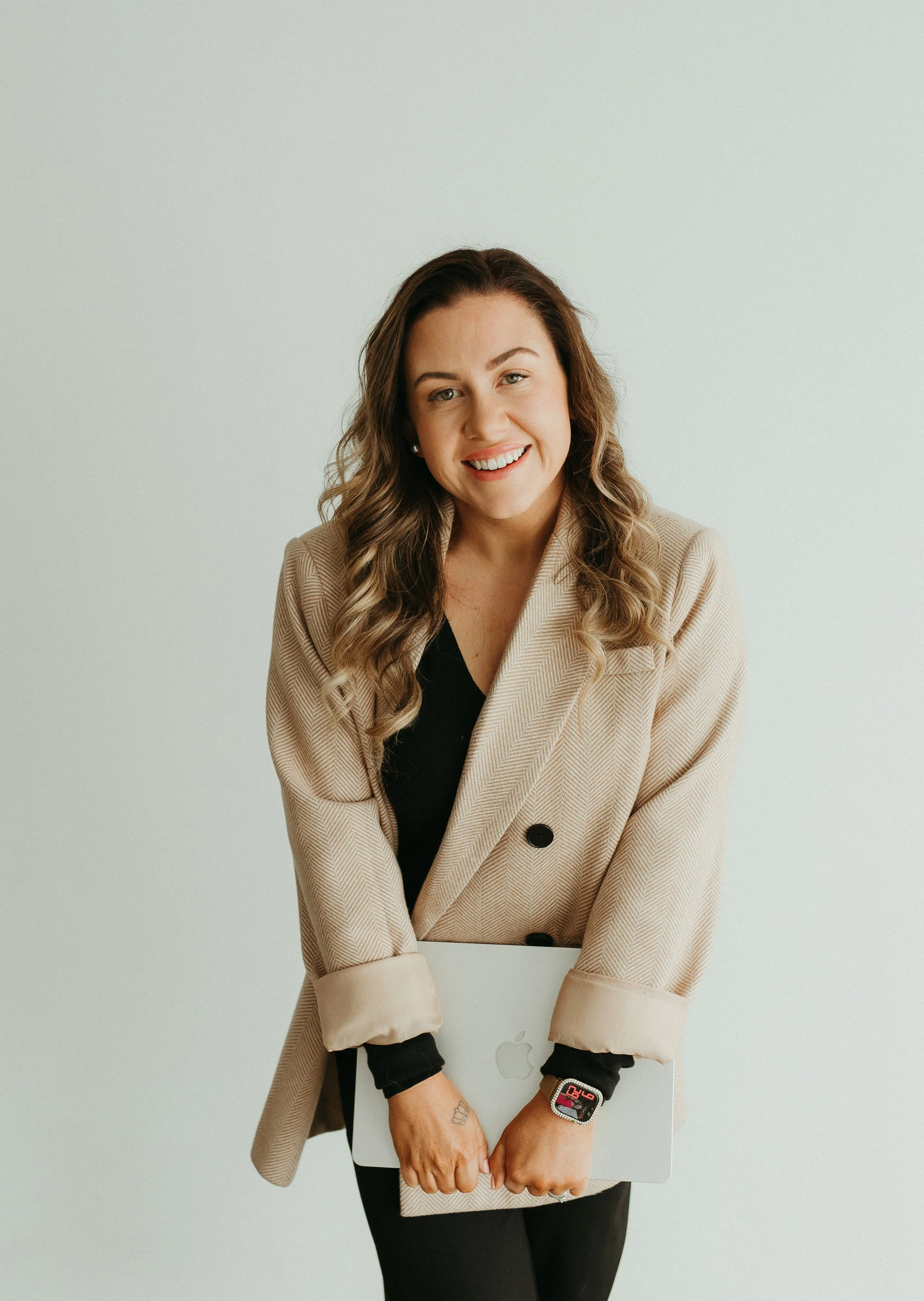 A woman with long, wavy brown hair, smiling, wearing a beige blazer with rolled-up sleeves, a black top, and a smartwatch, standing behind a silver laptop with an Apple logo on it, against a plain white background.