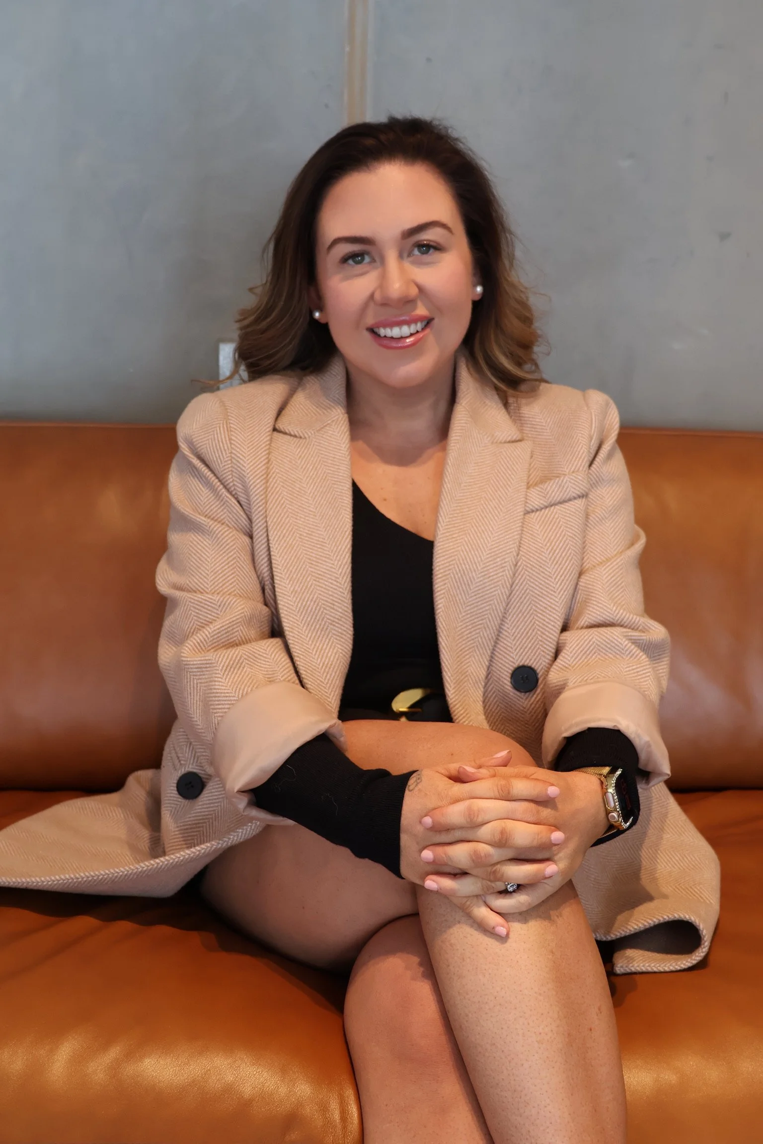 A woman with brown hair, wearing a beige blazer and black top, is sitting on a brown leather couch with her legs crossed, smiling at the camera, with a gray wall in the background.
