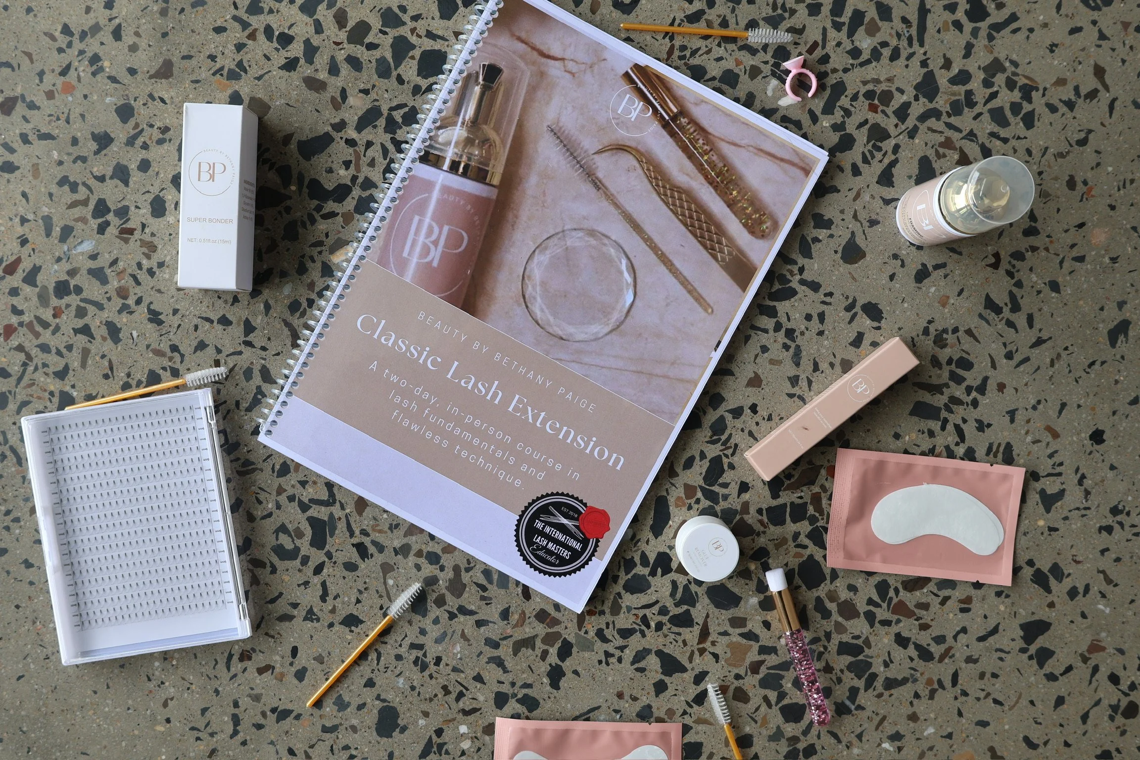 A flat lay of eyelash extension supplies including a booklet, a small bottle, a pink lash extension tray, a tray of individual eyelash extensions, pencils, and tools on a speckled terrazzo surface.