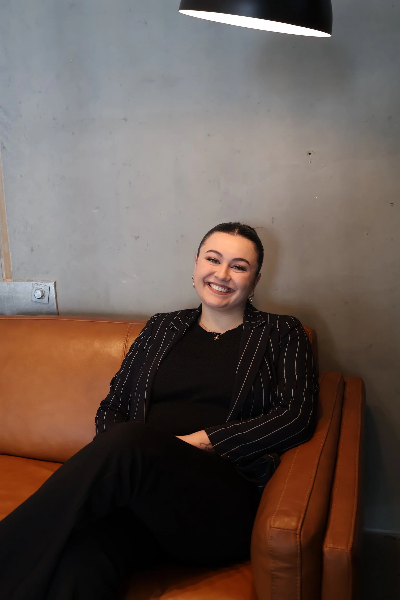 A smiling woman sitting on a brown leather couch against a gray wall, wearing a black top and a black and white striped blazer.