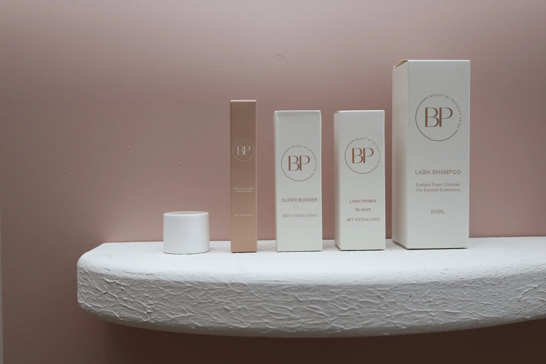 Display of four skincare or beauty product boxes, including lash shampoo, primer, super bonder, and a small container, arranged on a white textured shelf against a pink wall.