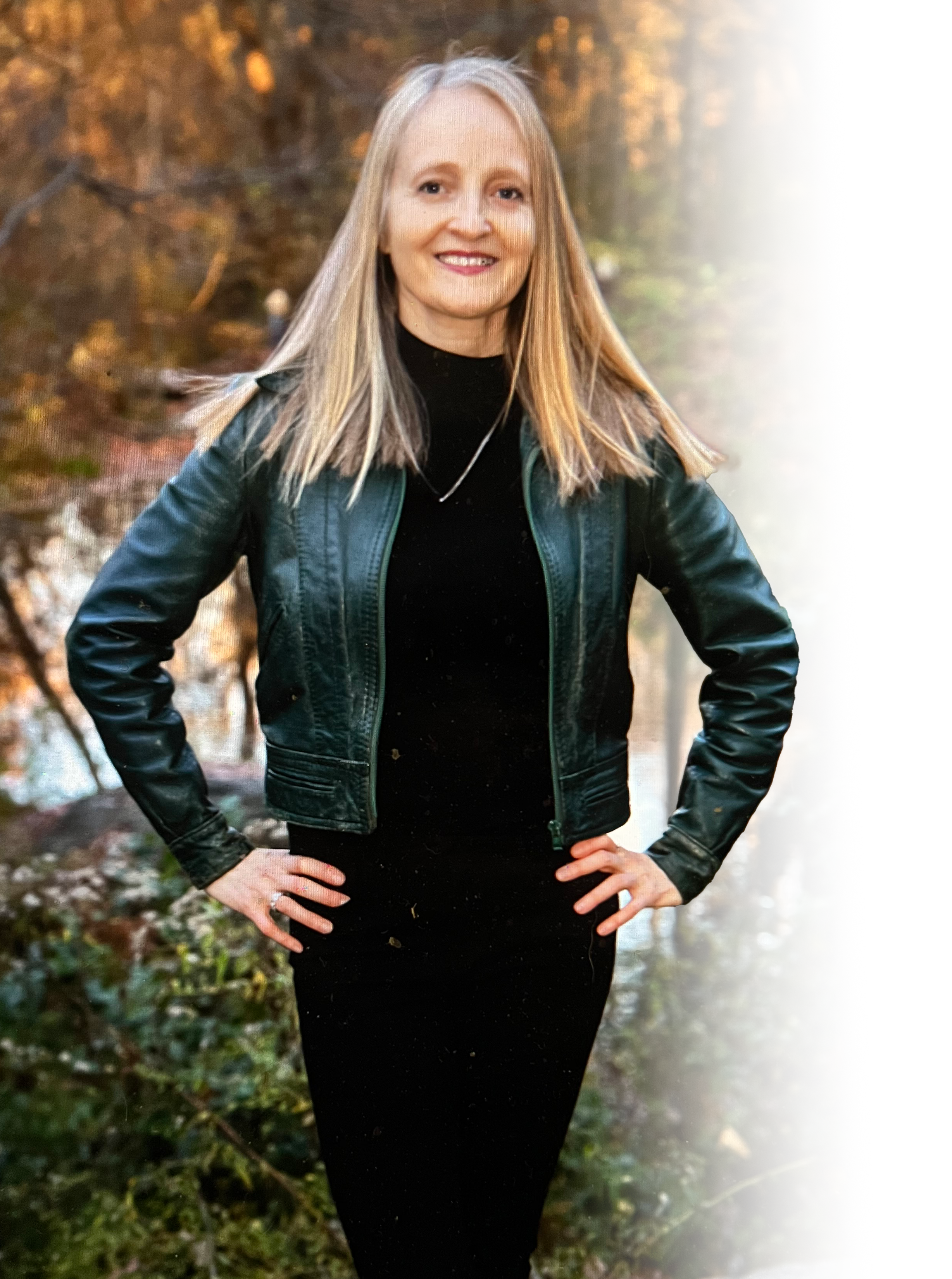 A woman with blonde hair smiling outdoors in autumn, wearing a black dress and a dark leather jacket, with hands on hips.