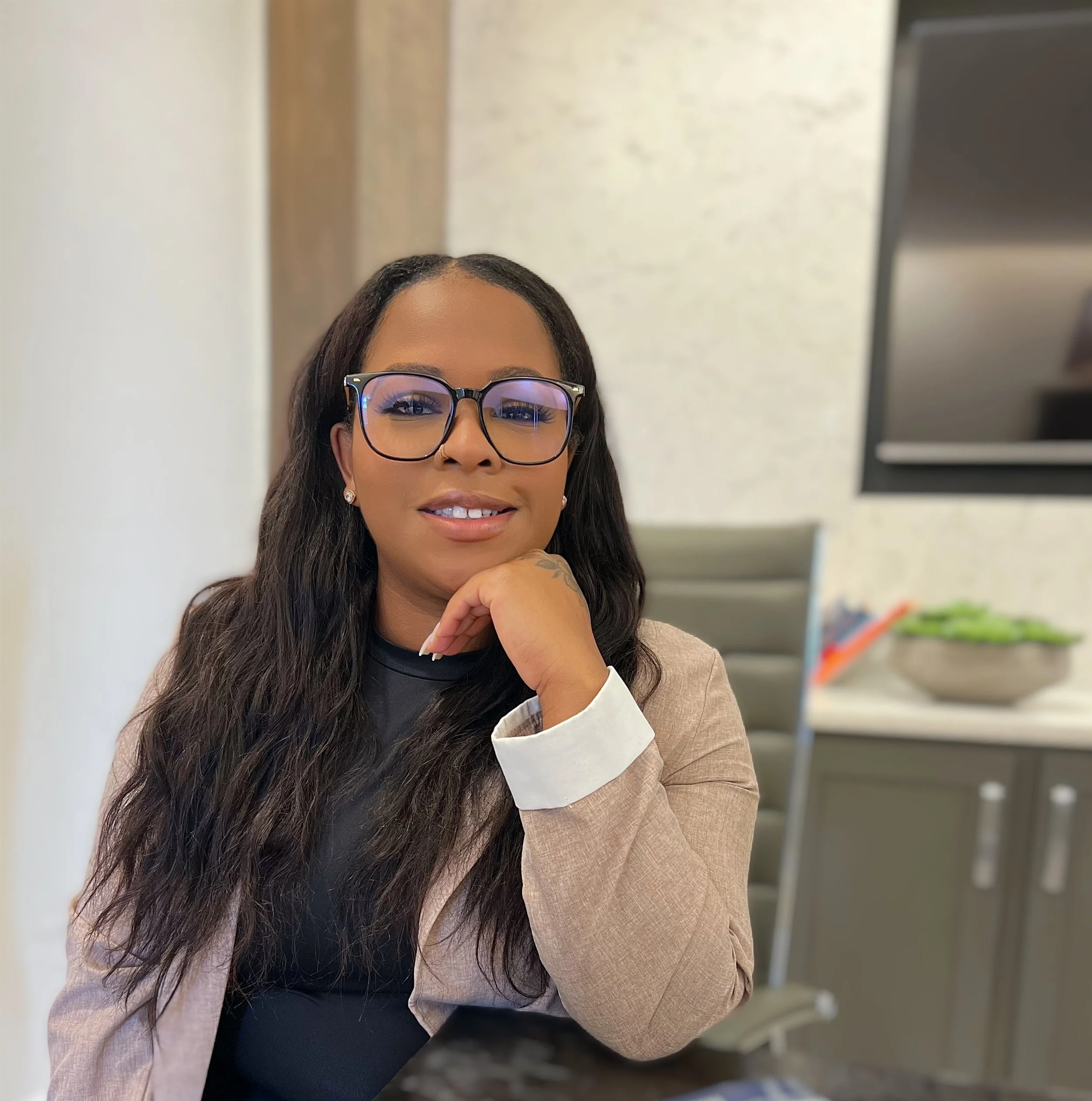 A woman wearing glasses and a beige blazer sitting at a desk in an office setting.