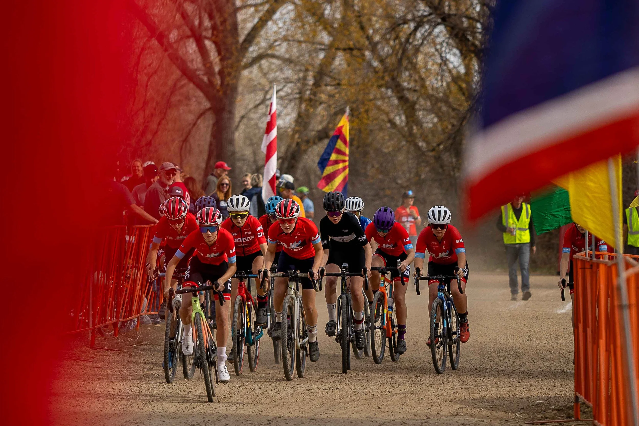 Cyclists participate in a BMX race on a dirt track lined with orange barriers, with spectators and flags in the background, surrounded by trees.