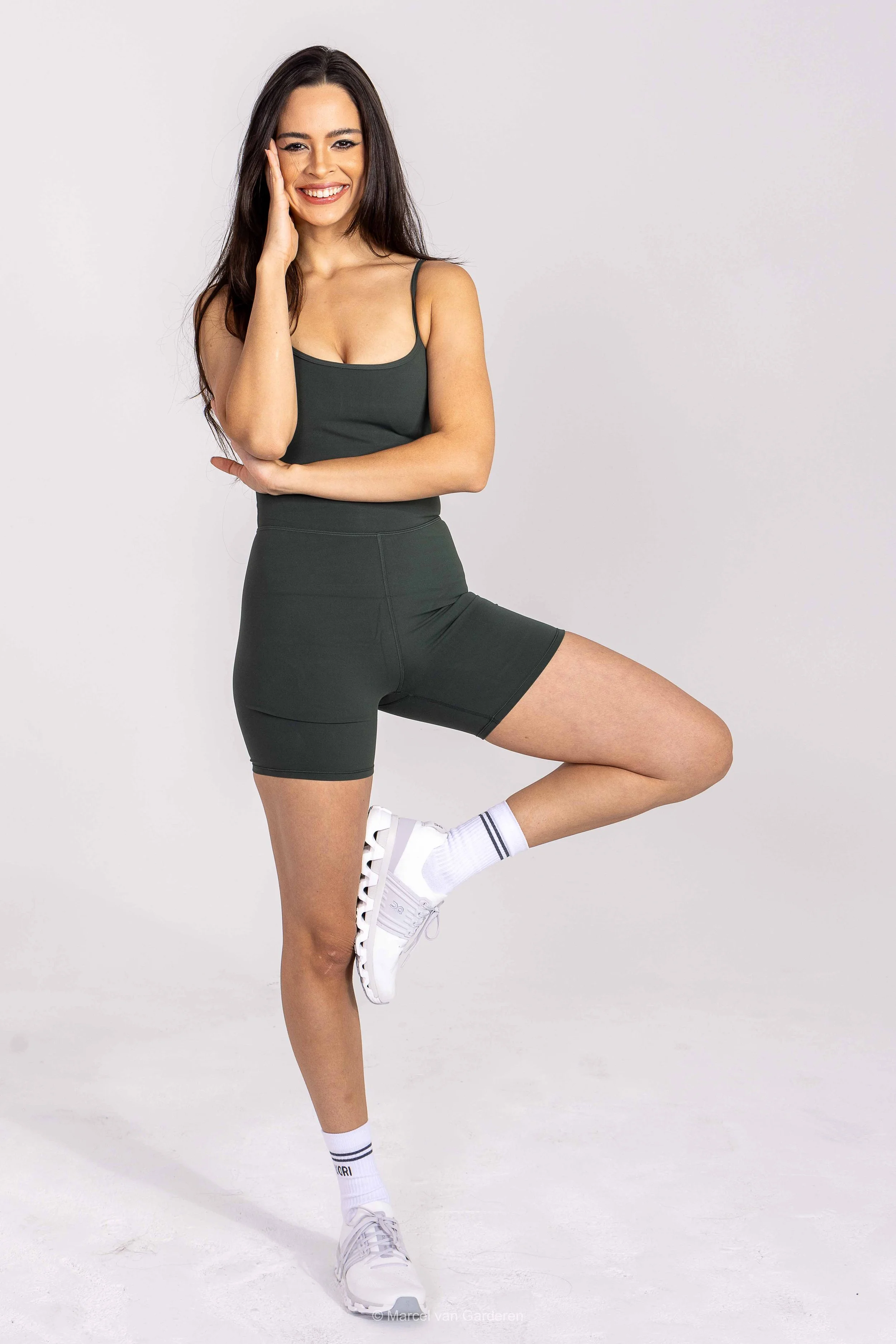 A young woman with long dark hair wearing a dark athletic outfit, white sneakers, and striped socks, standing in a fitness pose with one leg lifted and her hand on her face, smiling at the camera.