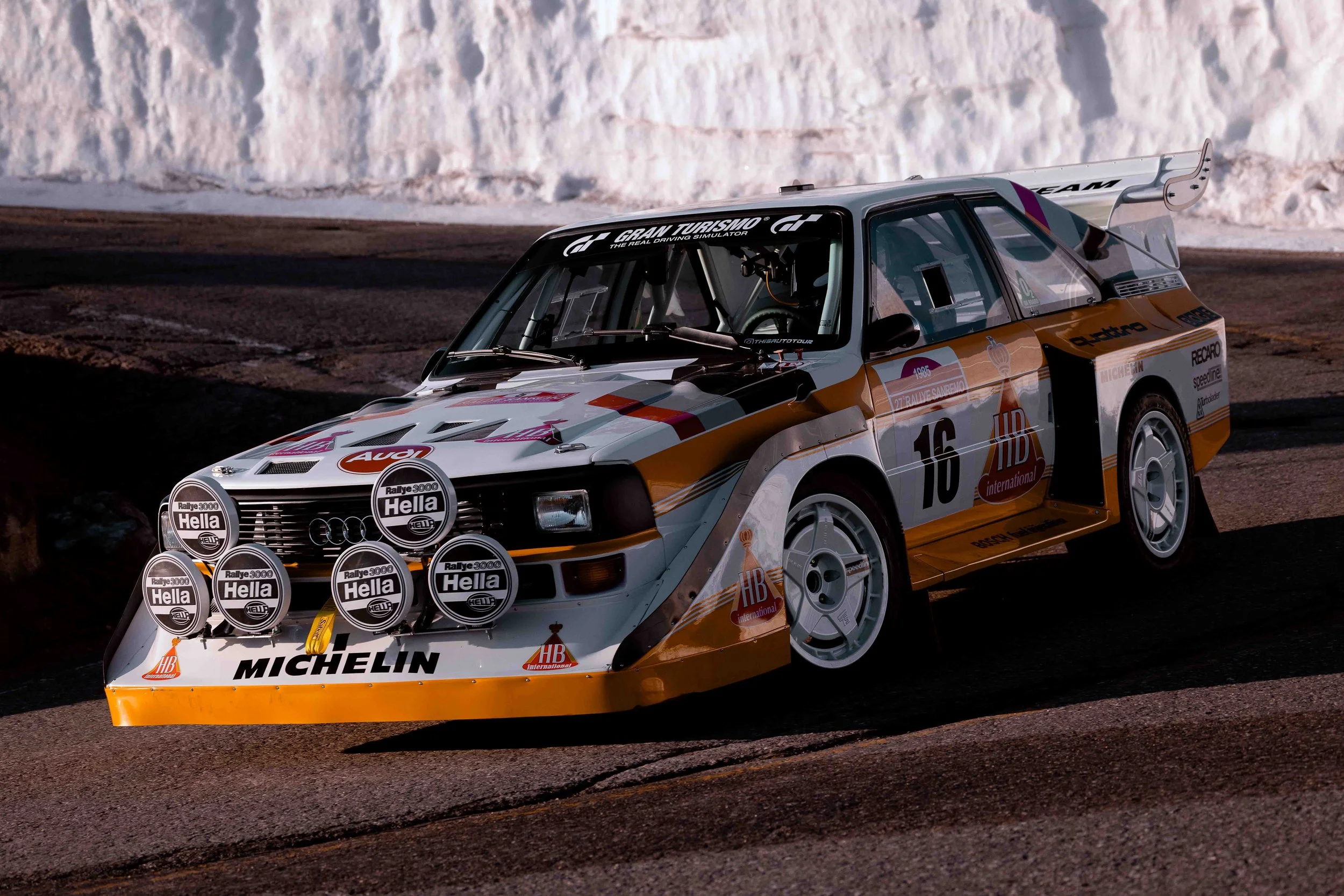 A vintage Audi rally car with racing decals and multiple front-mounted Hella lights, driving on a winding mountain road with snow-covered terrain in the background.