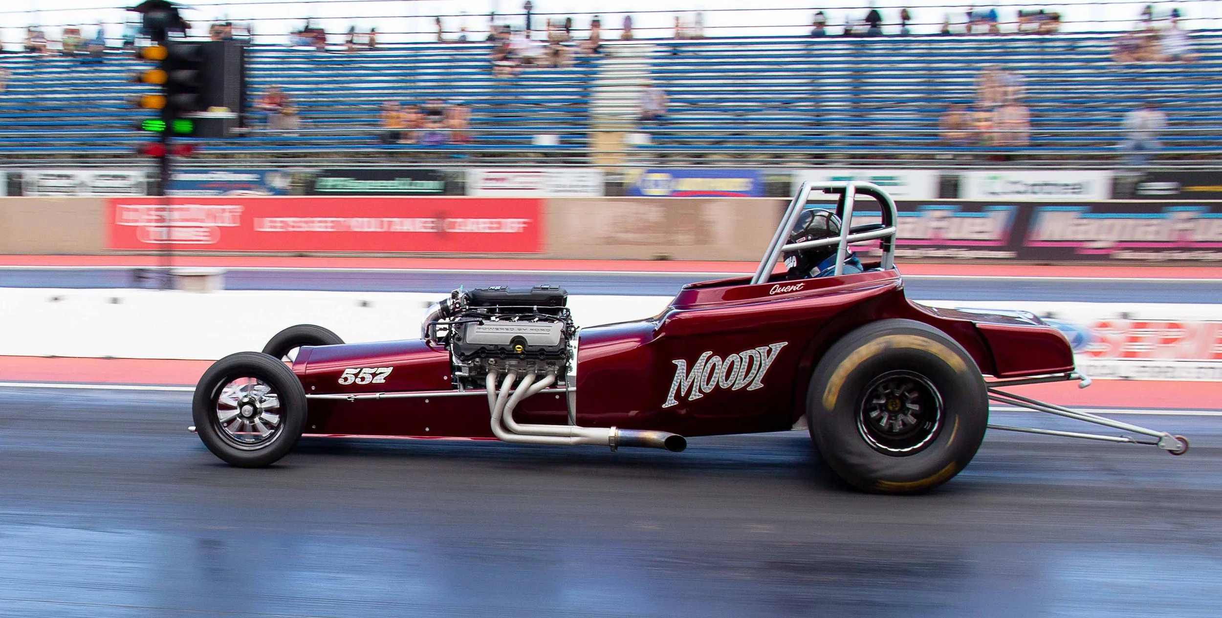 A vintage drag racing car, painted maroon with the name 'Moody' and the number '552', speeding down a drag strip.
