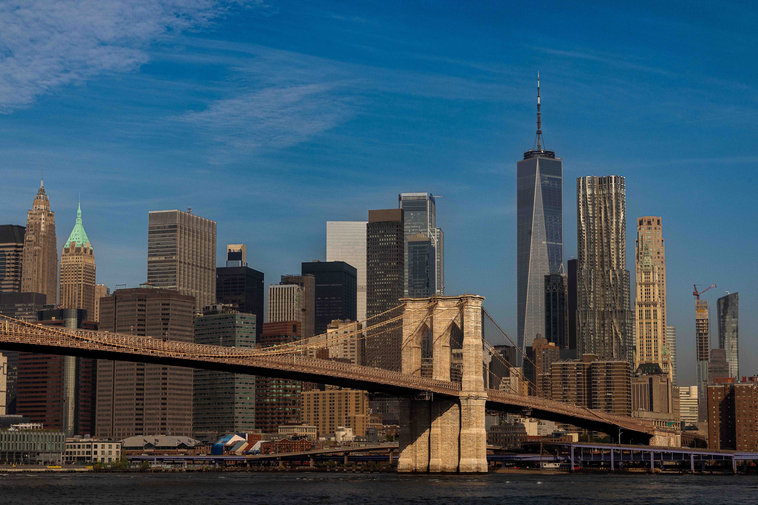 View of the New York City skyline with the Brooklyn Bridge in the foreground and various tall skyscrapers against a blue sky.