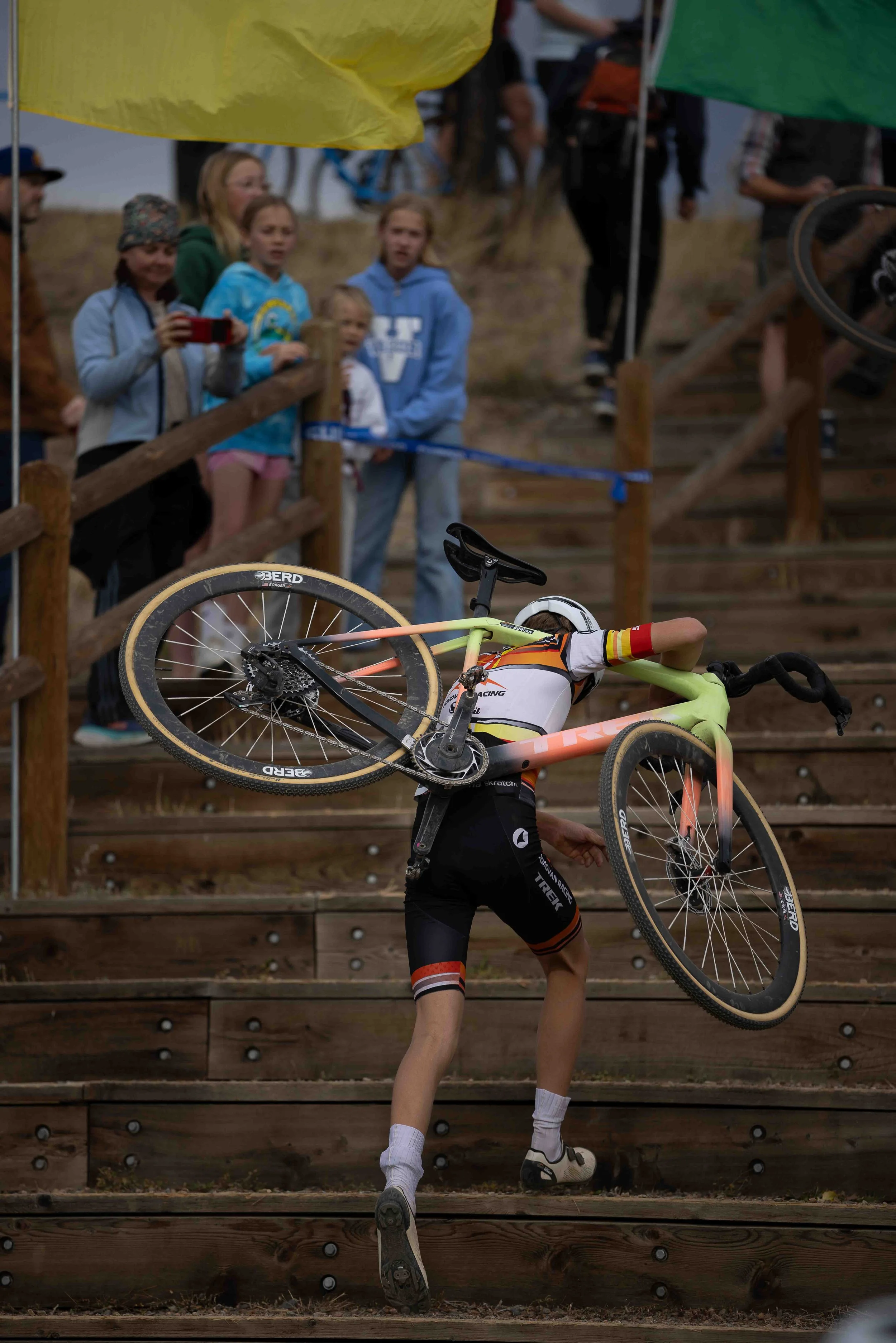 A cyclist carrying a bike on stairs during a competition, with spectators watching from the top of the stairs.