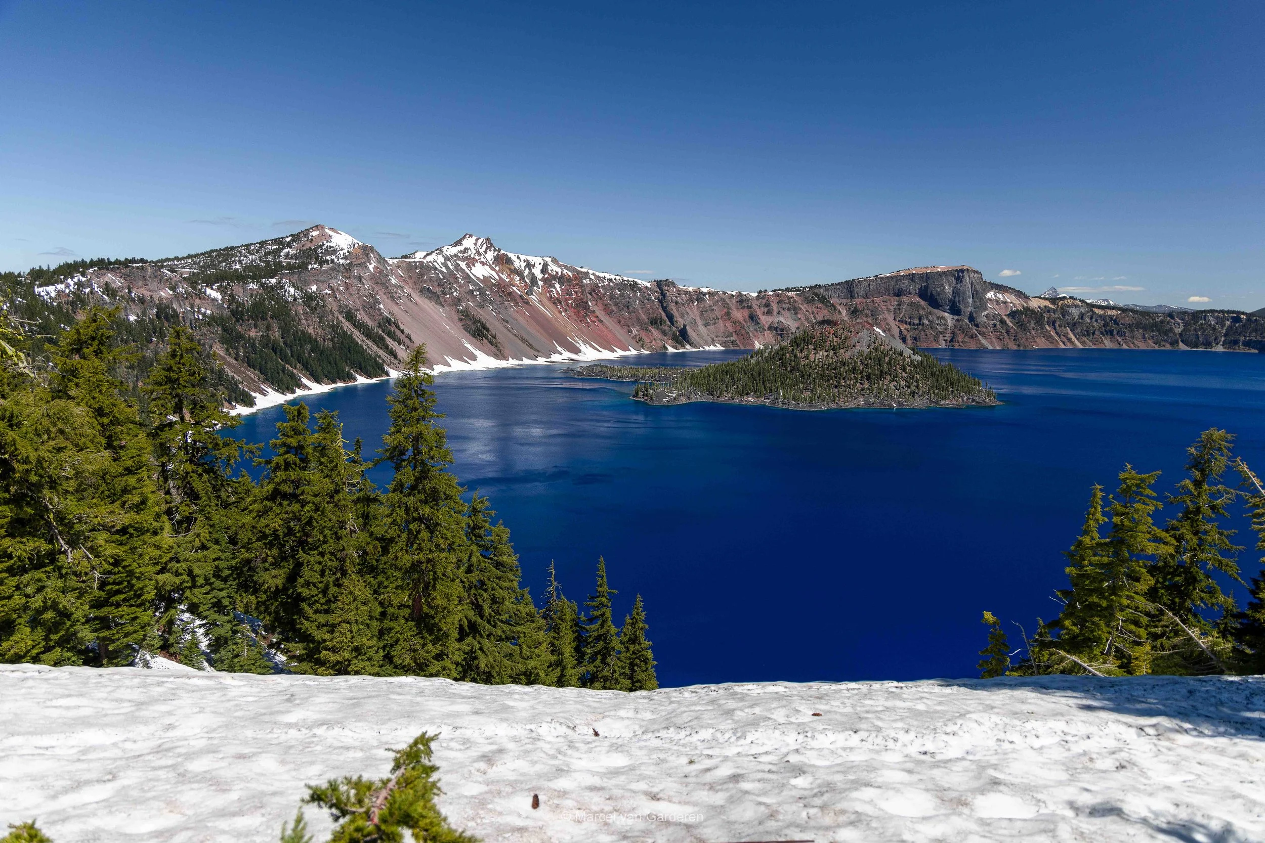 A scenic view of a deep blue lake surrounded by snow-capped mountains and green pine trees under a clear blue sky.